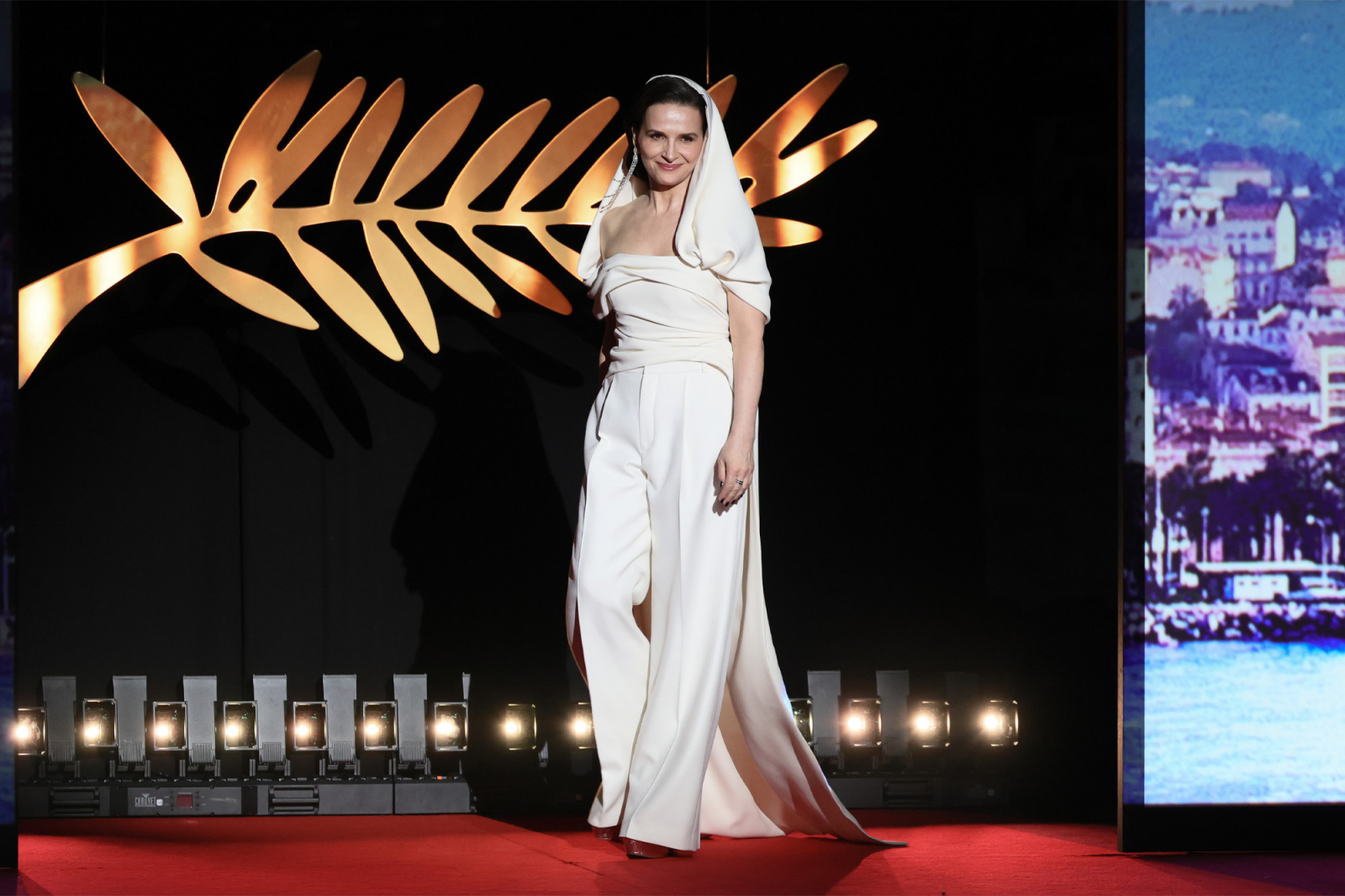 Juliette Binoche w białym stroju, składającym się z klasycznych spodni garniturowych i eleganckiej bluzki, spaceruje po czerwonym dywanie.