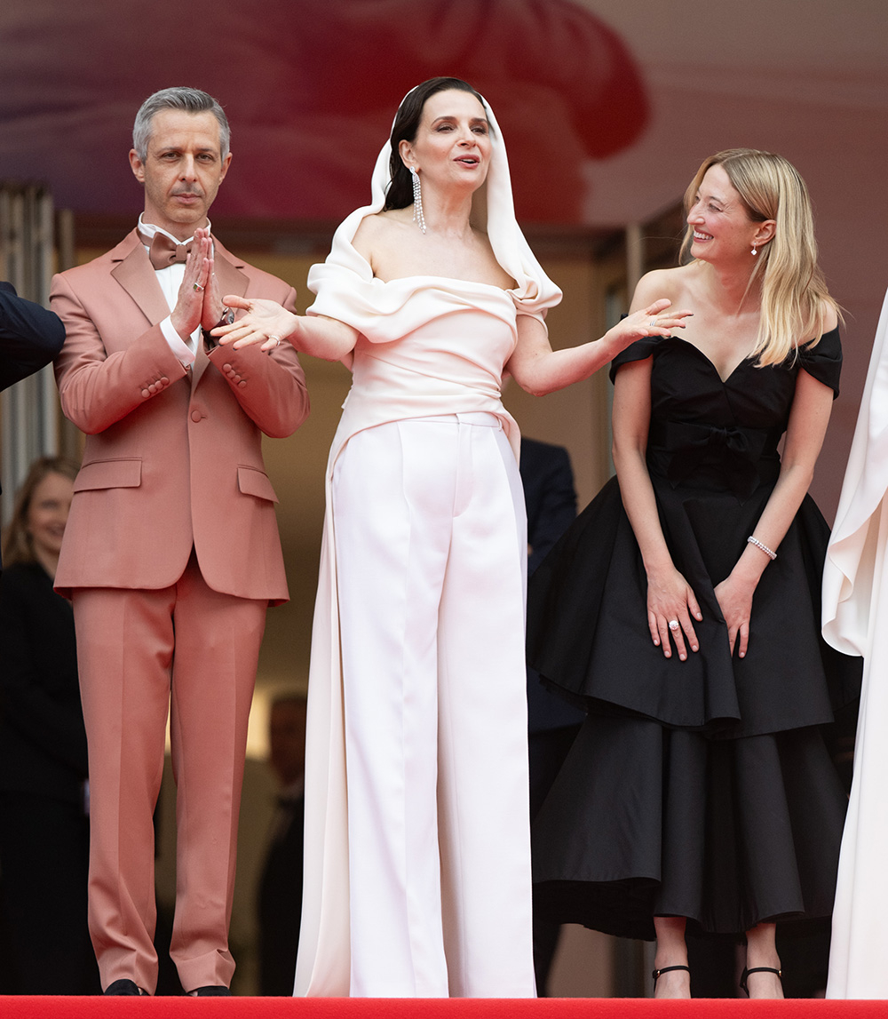 Na czerwonym dywanie stoją trzy osoby;Juliette Binoche ma na sobie klasyczne spodnie garniturowe w jasnym kolorze.
