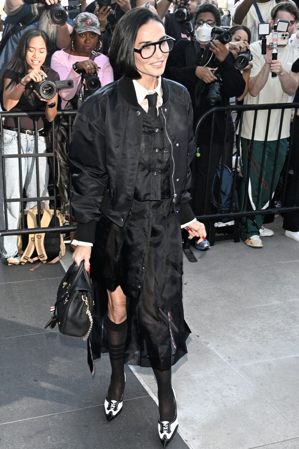 Demi Moore  w czarnej, eleganckiej stylizacji z bomberką, białą koszulą i długą sp&oacute;dnicą, przechodzi obok grupy fotograf&oacute;w.