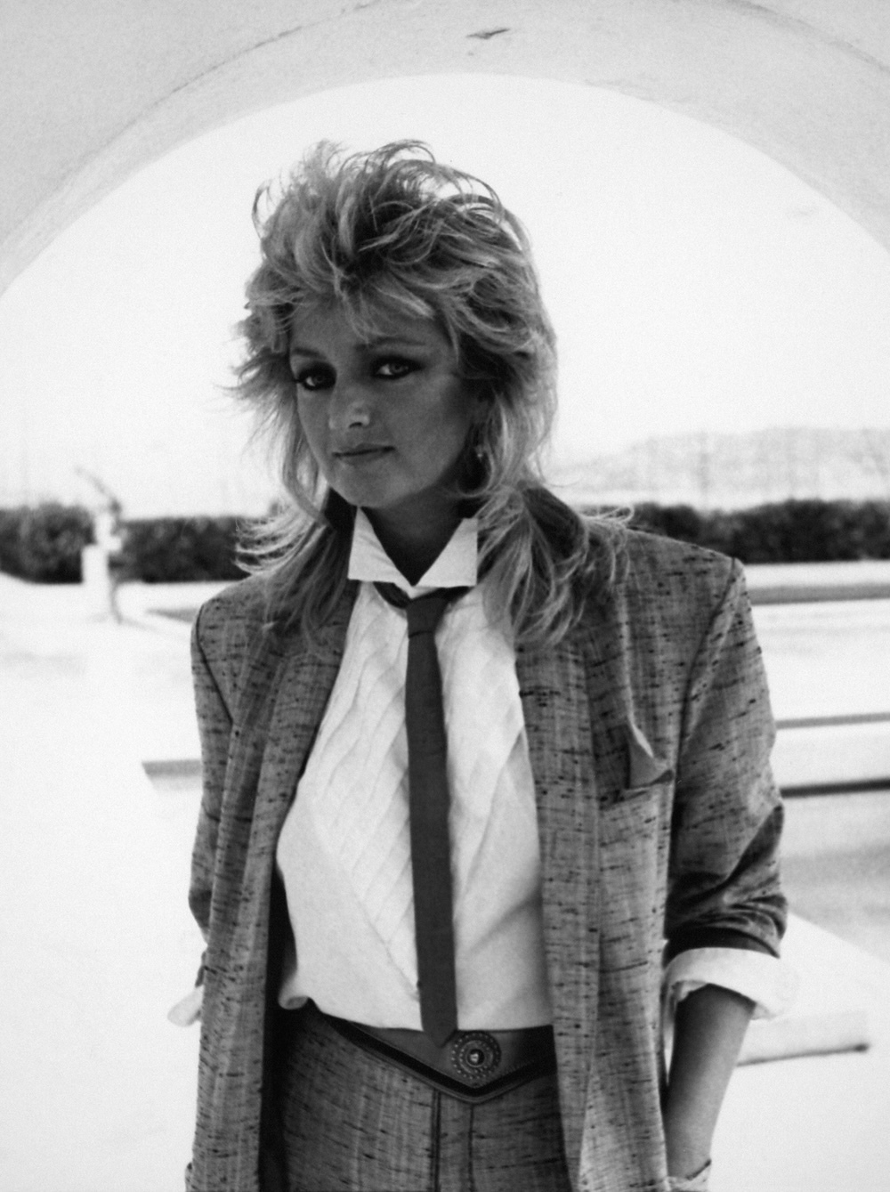 Bonnie Tyler z długimi włosami w eleganckim, szarym garniturze z krawatem, stoi na zewnątrz w jasnym otoczeniu.
