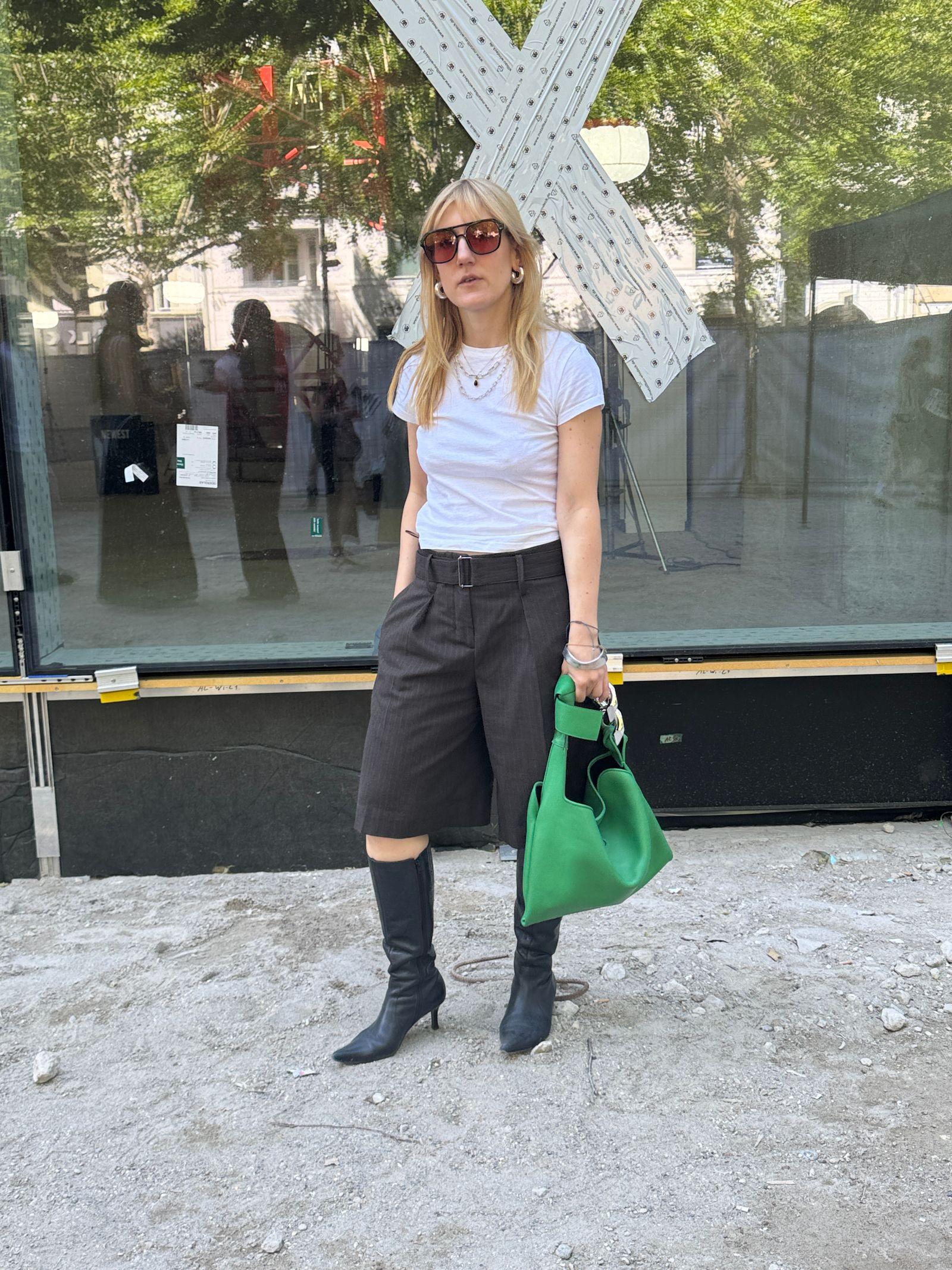 Kobieta w białym t-shircie, czarnych szortach bermudach, czarnych botkach, trzyma zieloną torebkę. Tydzień mody w Berlinie.