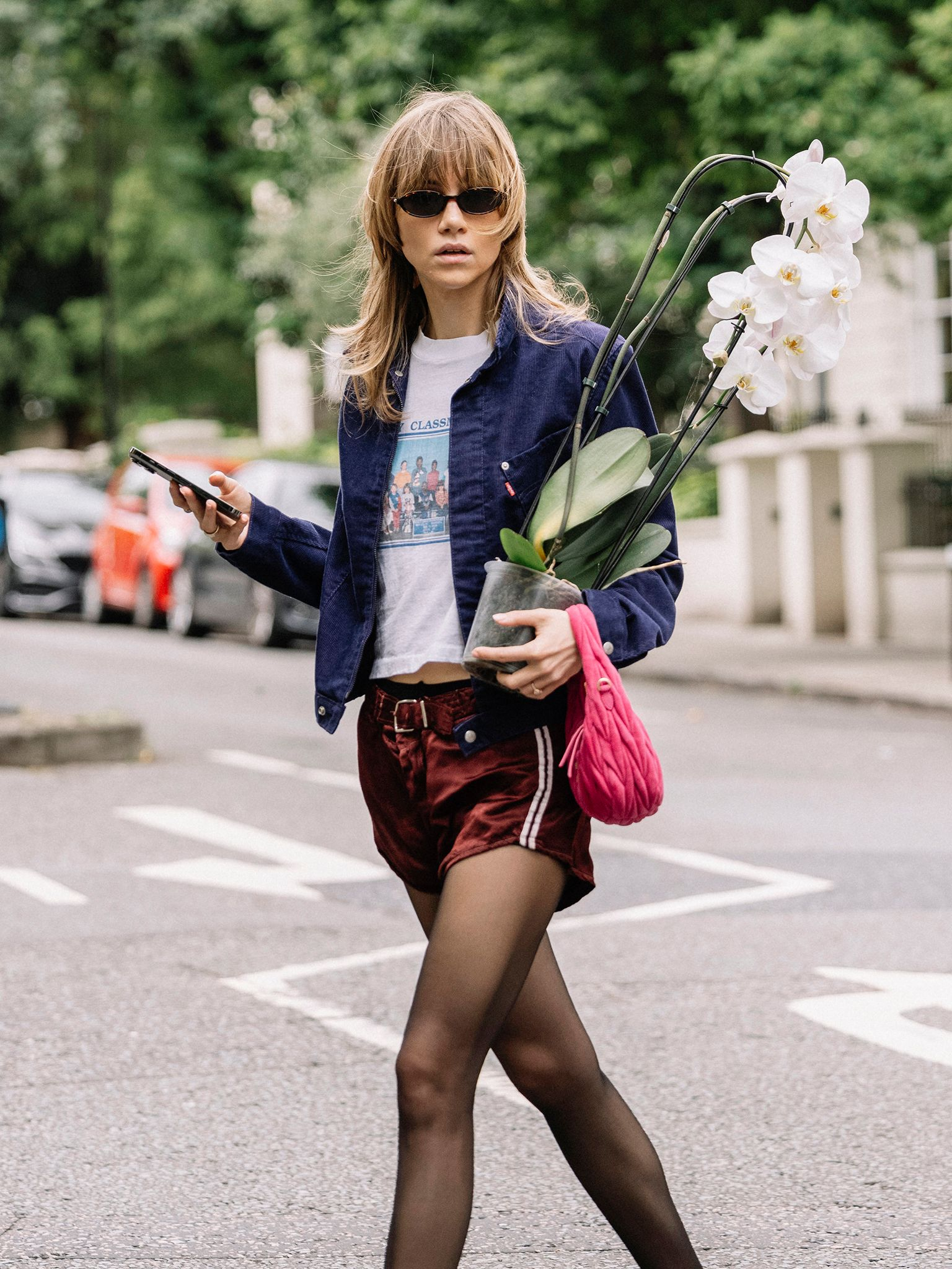 Suki Waterhouse w bordowych sportowych szortach, t-shircie z nadrukiem i kurtce, trzyma doniczkę z orchideami oraz telefon.