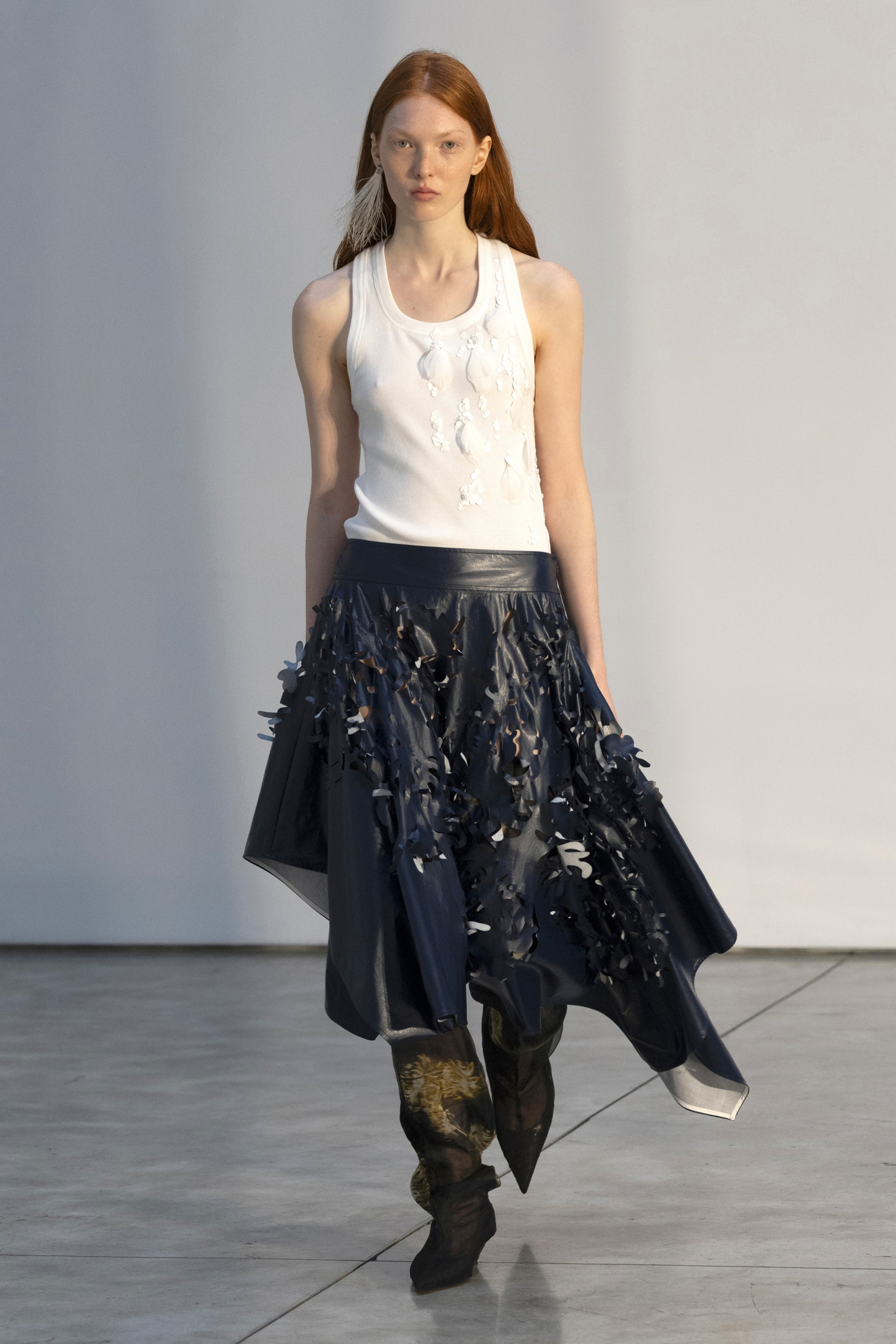 Modelka w asymetrycznej spódnicy midi i białym topie, prezentująca kreację Proenza Schouler.