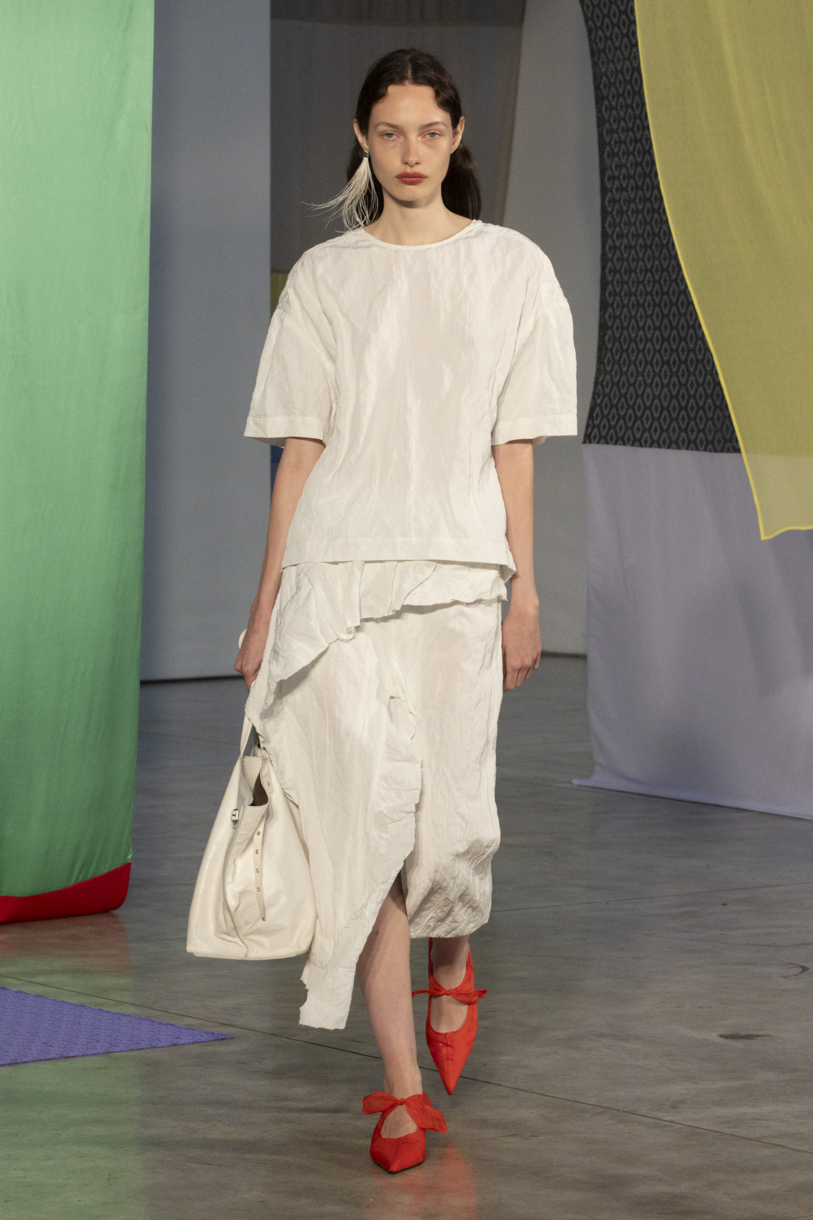 Modelka prezentuje kreację Proenza Schouler, składającą się z białej spódnicy midi i czerwonych butów.