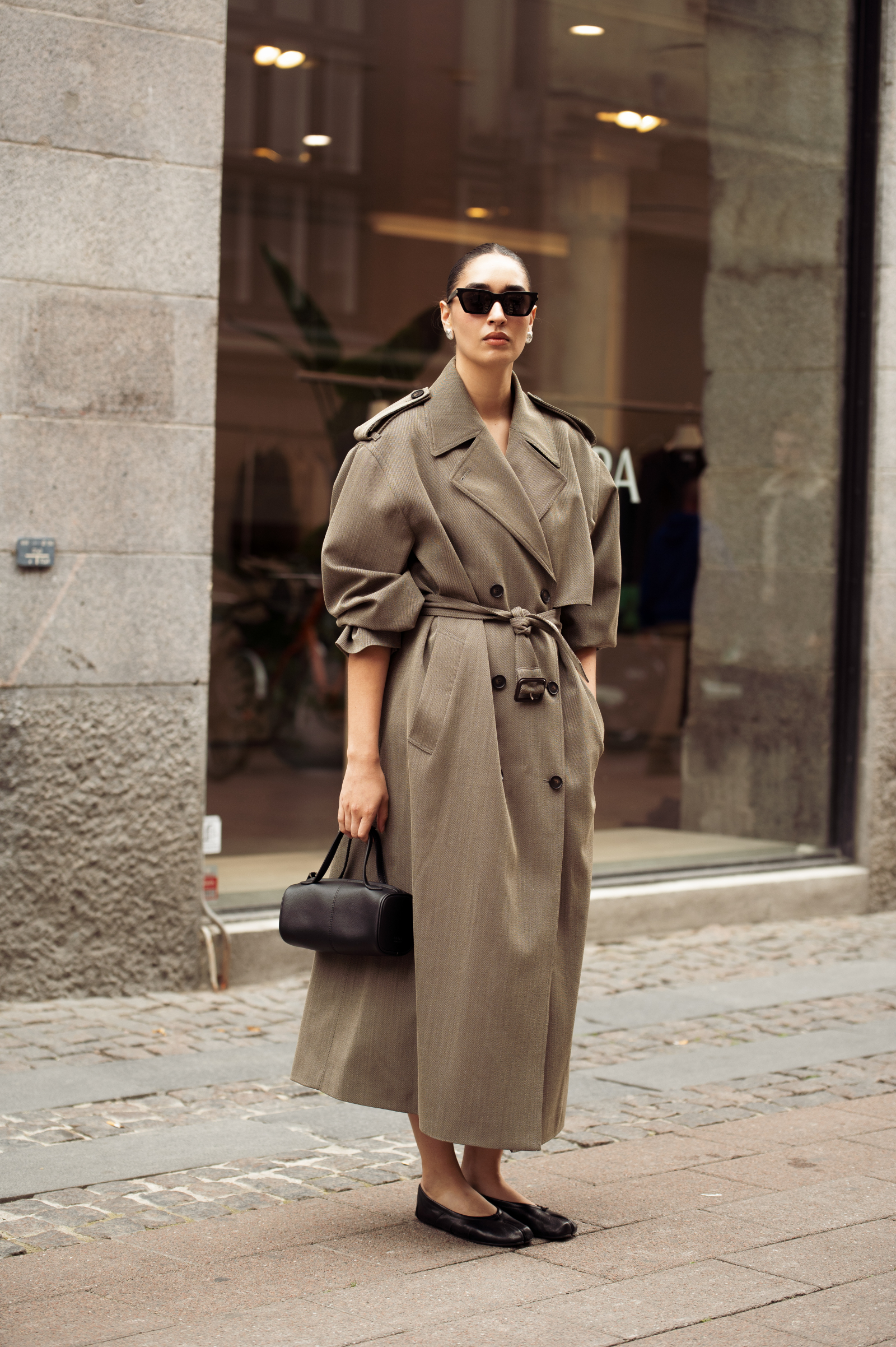 Stylowa osoba w trenczu oversize, nosząca baleriny i torebkę, stoi na miejskiej ulicy.