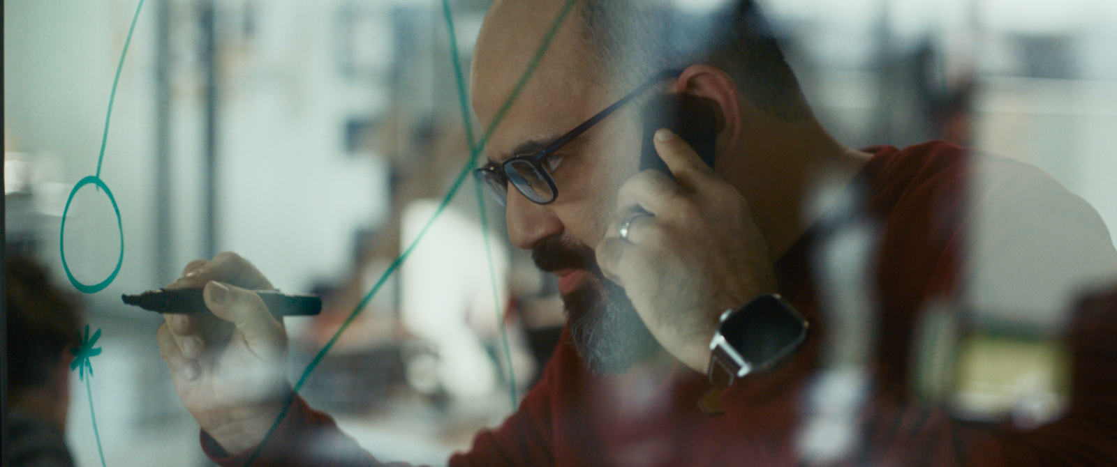 Mężczyzna z brodą rozmawia przez telefon i szkicuje coś na przezroczystej powierzchni, głos hind rajab w kadrze.