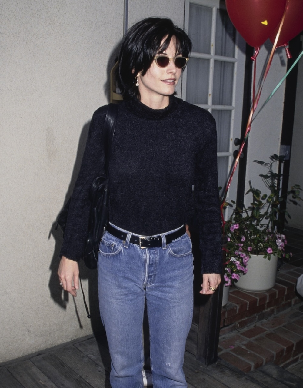 Courtney Cox w czarnym swetrze i jeansach damskich z 1995 roku, przy wejściu z balonami w tle.