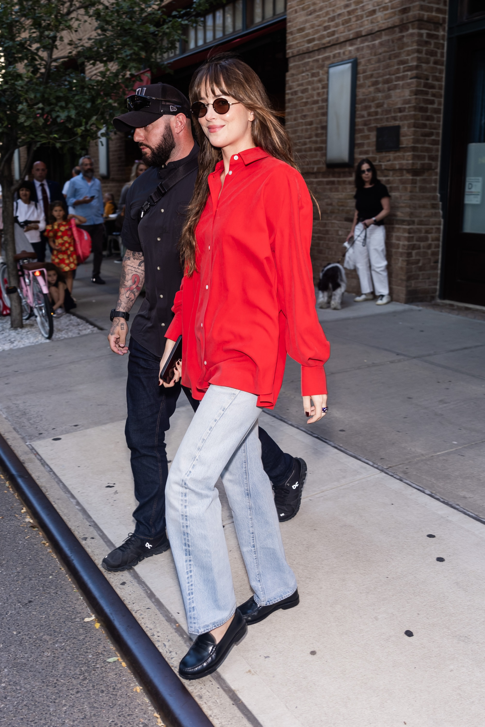 Dakota Johnson w czerwonej koszuli damskiej, jasnych jeansach i czarnych mokasynach spaceruje z towarzyszem.