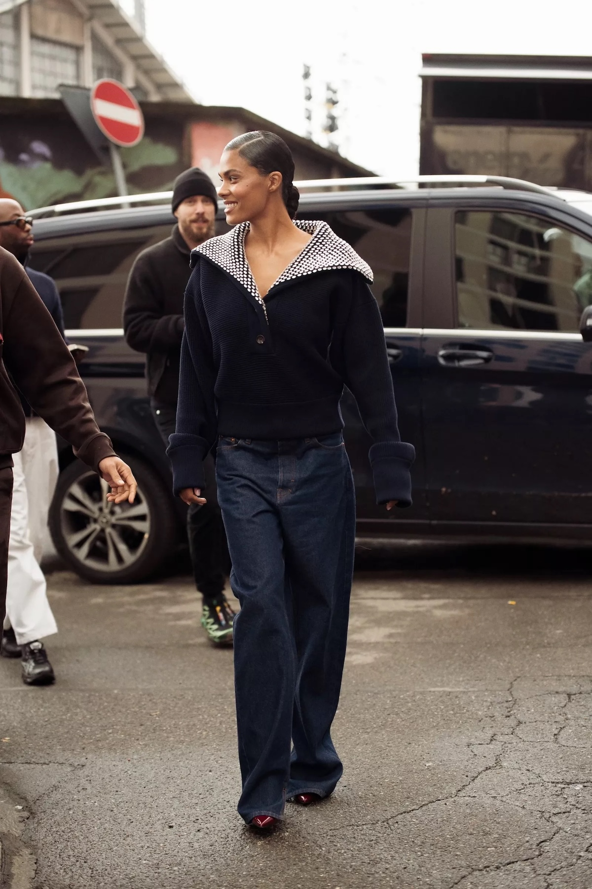 Tina Kunakey w granatowych jeansach damskich i bordowych szpilkach spaceruje po ulicy obok innych osób.