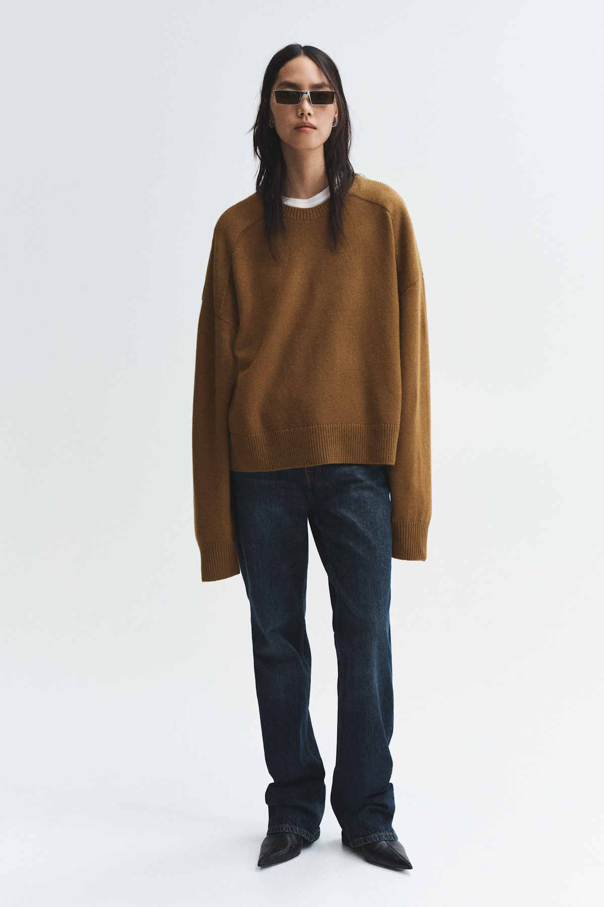 Modelka w brązowym swetrze, ciemnoniebieskich dżinsach i eleganckich butach na obcasie, z okularami przeciwsłonecznymi. Kolekcja H&M Studio Essentials na sezon jesień-zima 2025.