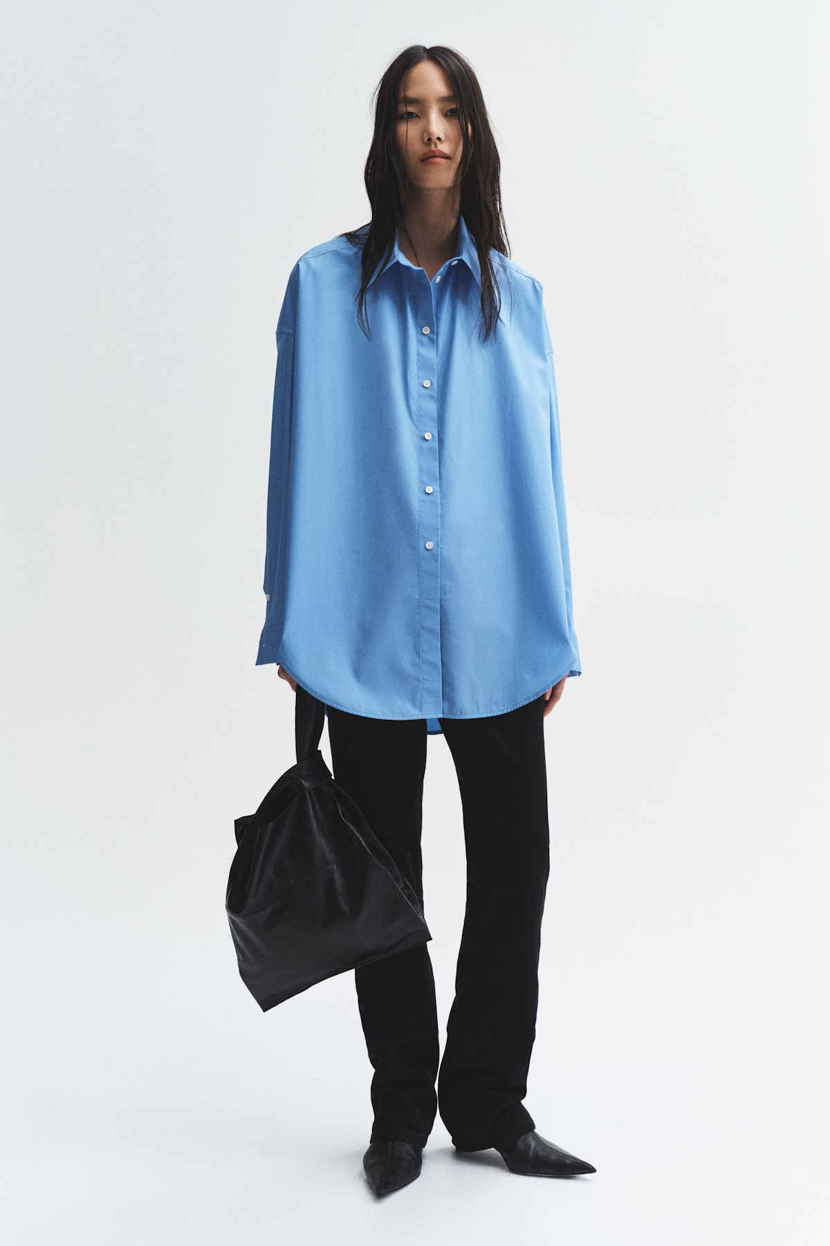Modelka w niebieskiej, dużej koszuli oraz czarnych spodniach trzyma torbę w kolorze czarnym. Tło jasne i minimalistyczne. Kolekcja H&M Studio Essentials na sezon jesień-zima 2025.