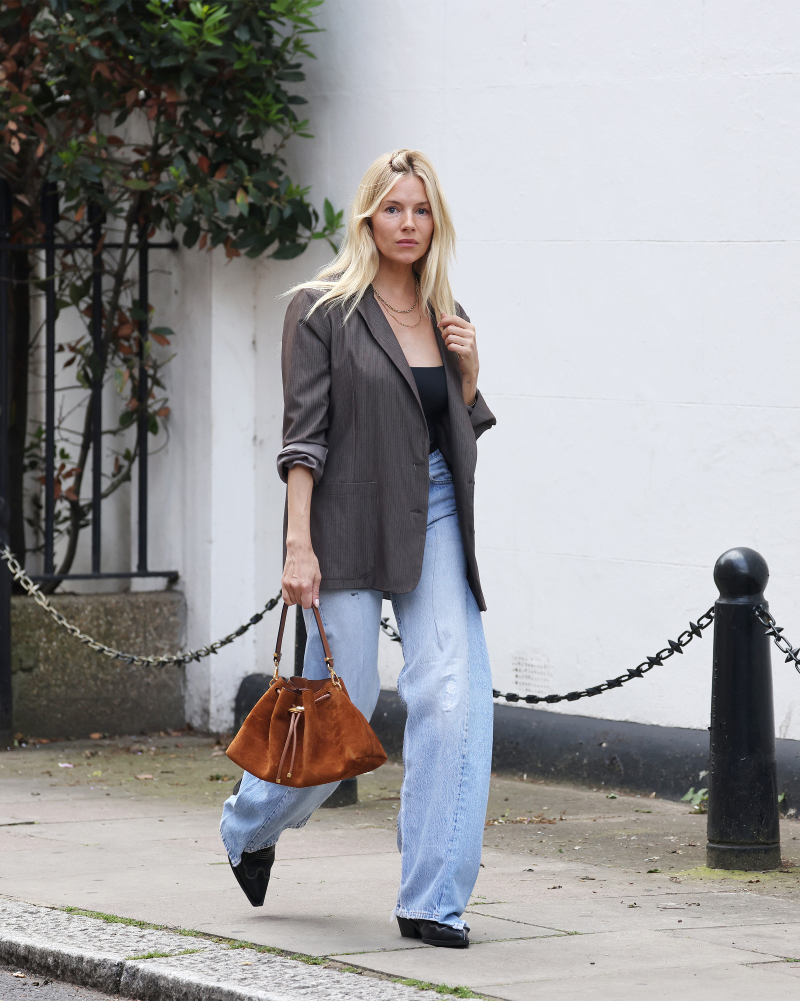 Sienna Miller w błękitnych jeansach barrel z wysokim stanem oraz szarym żakiecie, idzie po ulicy z torbą.