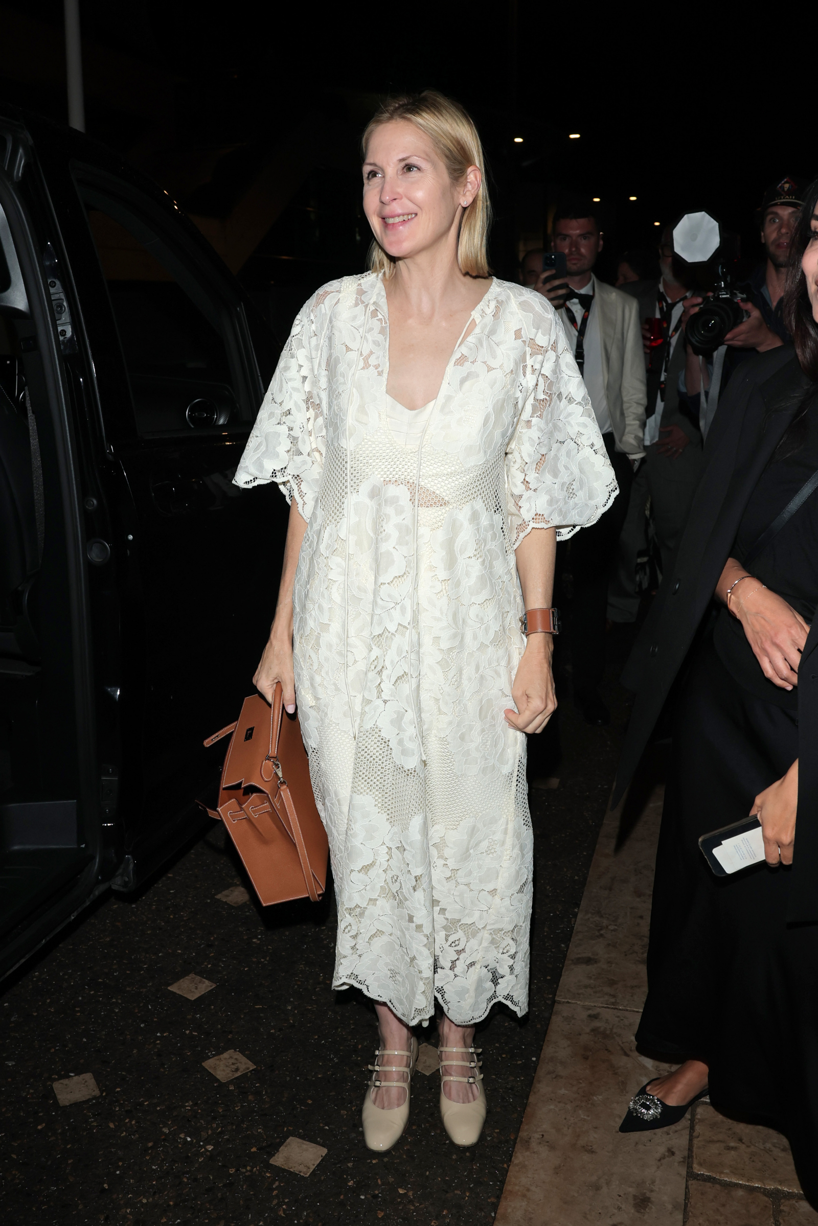 Kelly Rutherford w białej sukience koronkowej boho, z brązową torebką, uśmiechnięta, przy samochodzie.