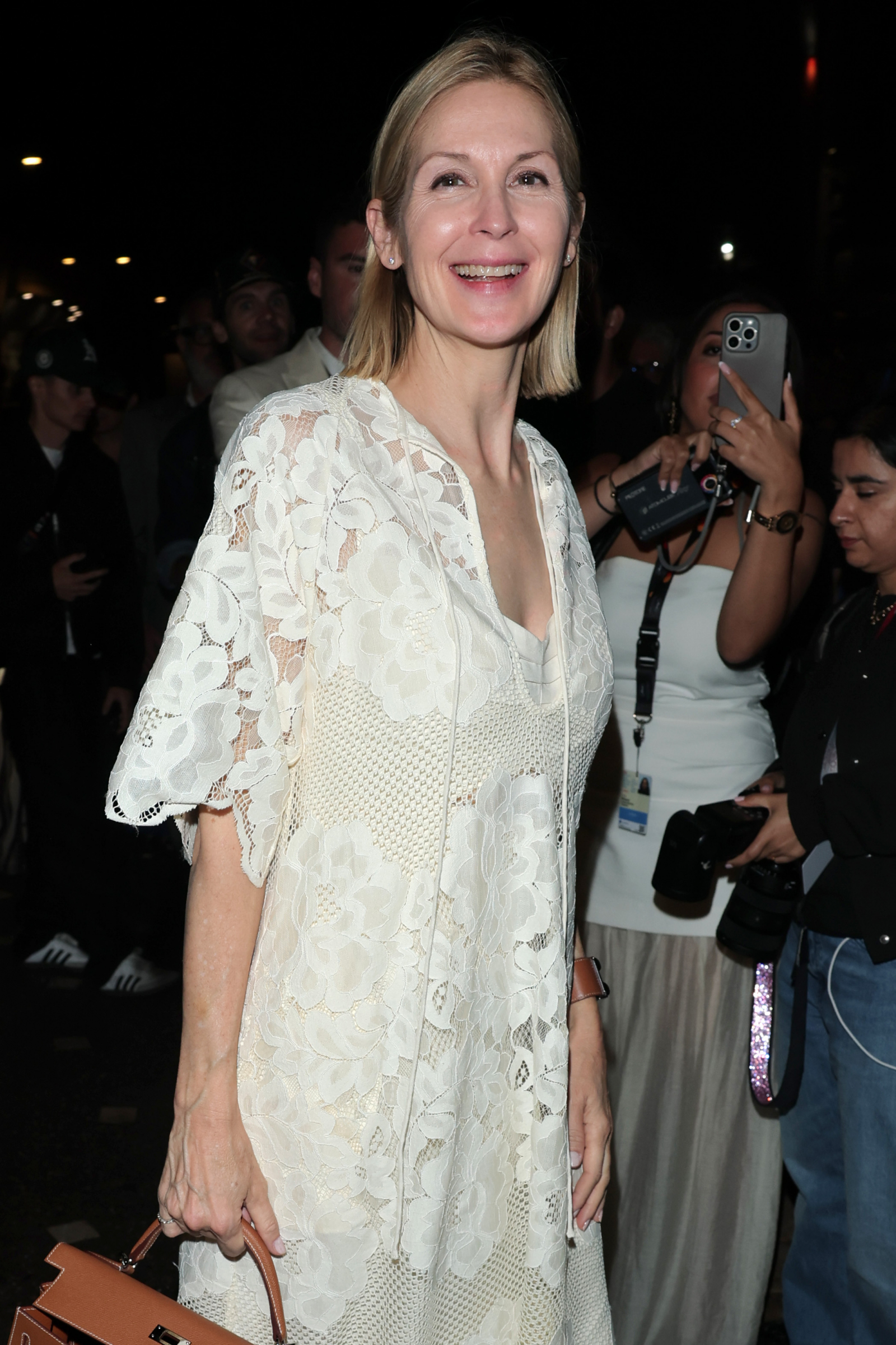 Kelly Rutherford w białej sukience koronkowej boho, uśmiechnięta, otoczona przez innych ludzi w tle.