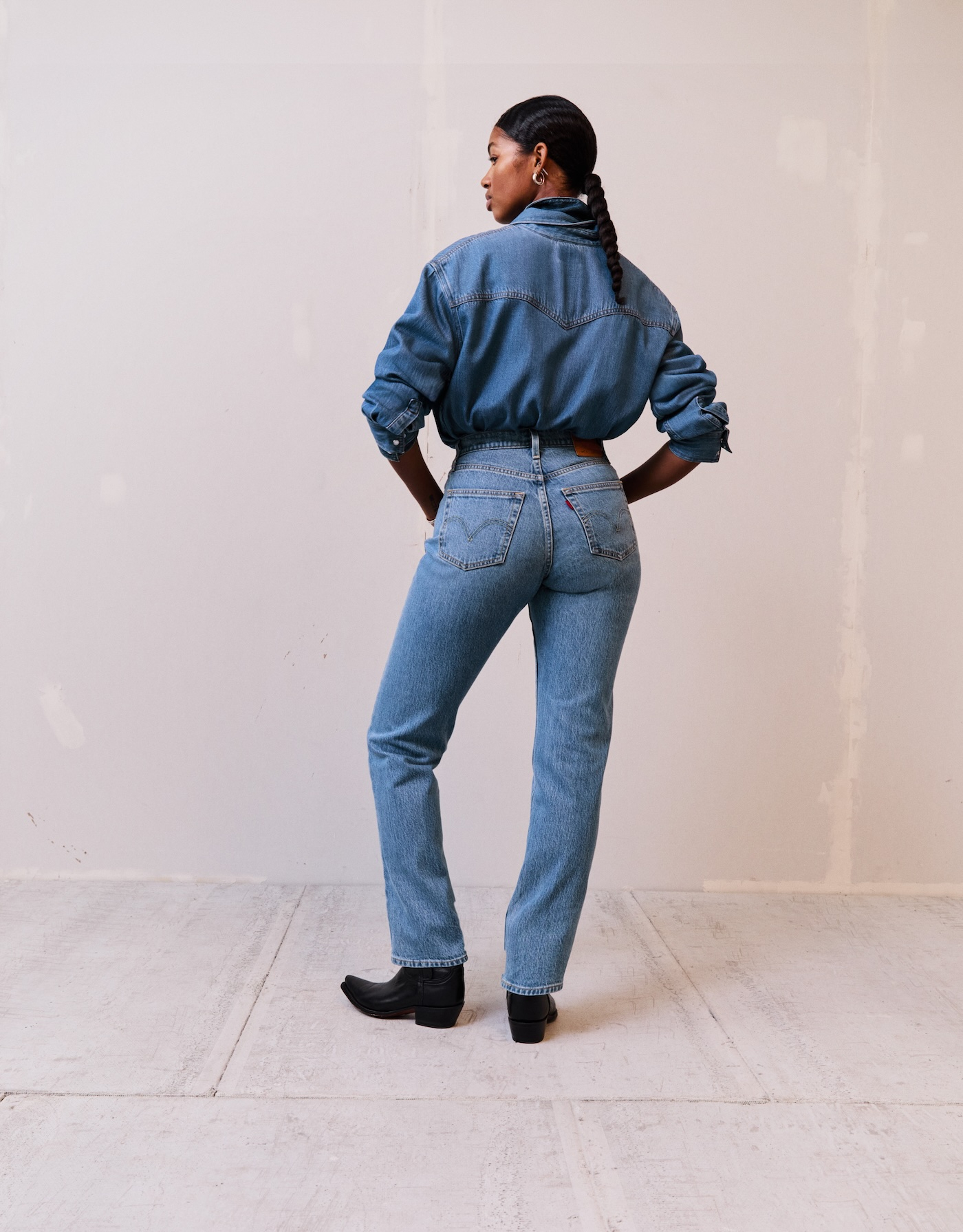 Modelka w jeansach damskich Levi's 501 Curve, jeansowej koszuli i czarnych butach kowbojkach pozuje na białym tle.