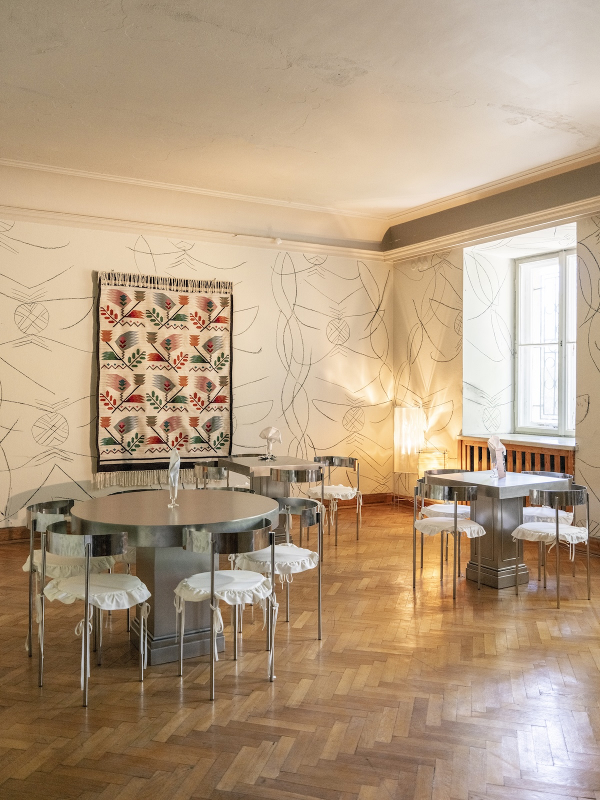 Wnętrza Lupo Aperitivo Bar w Willi Gawrońskich, gdzie metalowe stoliki otaczają kilim według archiwalnego projektu Zofii Stryjeńskiej, a ściany zdobią rysunki Martyny Wojtuszek.