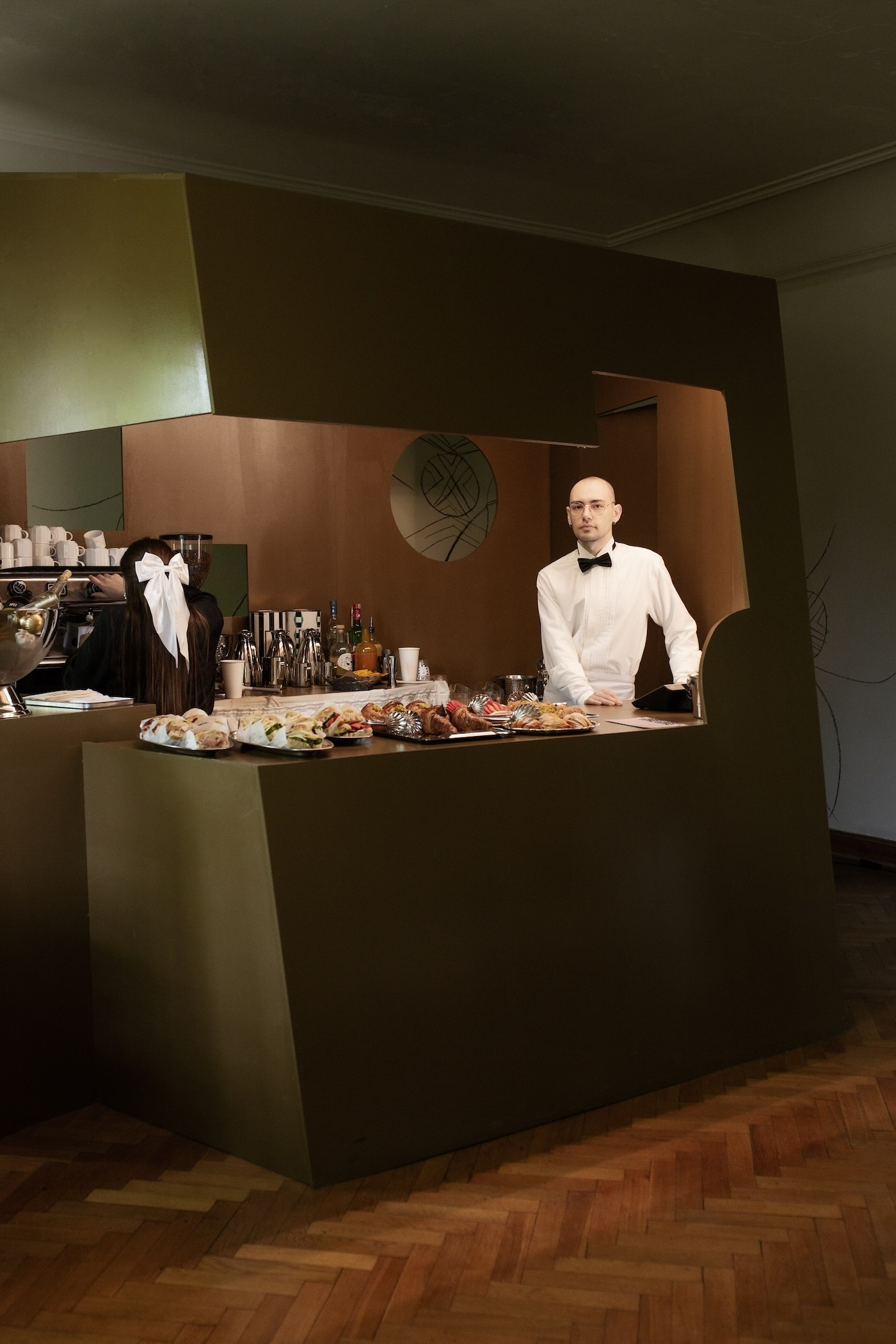 Wnętrza Lupo Aperitivo Bar w Willi Gawrońskich, widoczny bar w kolorze oliwkowym, za barem dwie kelner i kelnerka, ekspres do kawy, słodkości i kanapki.