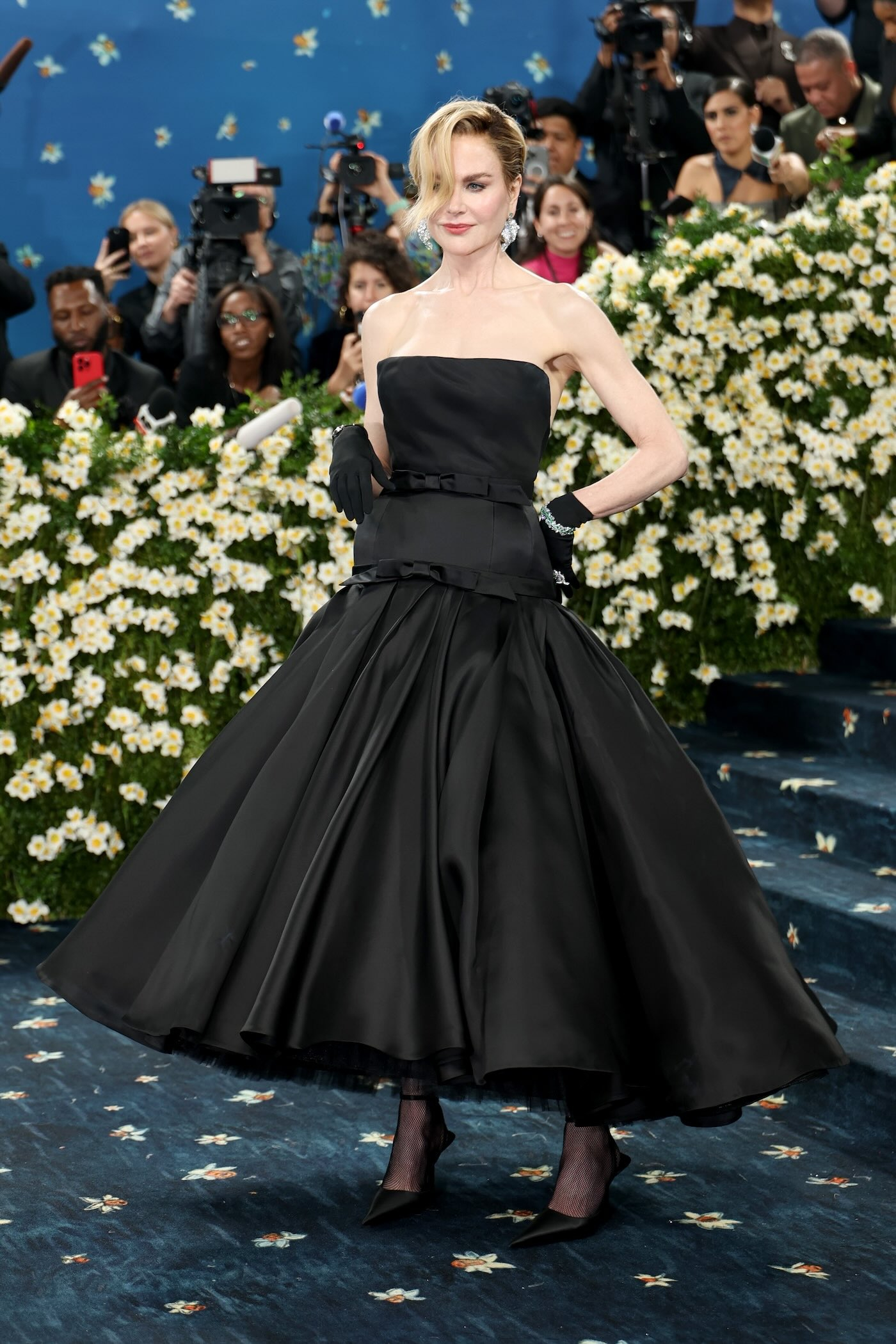 Nicole Kidman podczas MET Gala 2025 ma na sobie czarną gorsetową suknię wieczorową, z rozkloszowanym dołem, licznymi halkami, satynowymi wykończeniami w kształcie kokardek, do tego nosi czarne rękawiczki oraz szpilki.
