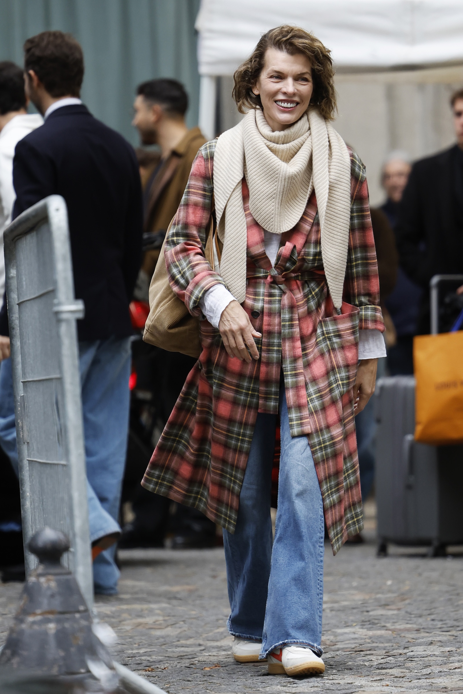 Milla Jovovich w szerokich jeansach damskich i białych sneakersach z uśmiechem spaceruje w miejskim otoczeniu.
