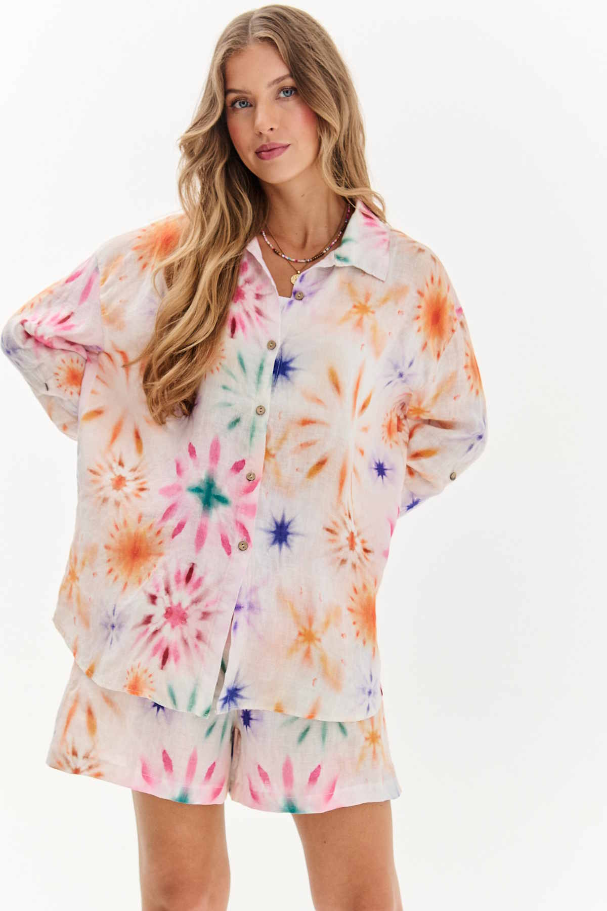 Kobieta w luźnej, kolorowej koszuli i szortach z wzorem tie-dye, uśmiechnięta, z włosami falowanymi. Kolekcja Sunspell polskiej marki Naoko na lato 2025 roku.