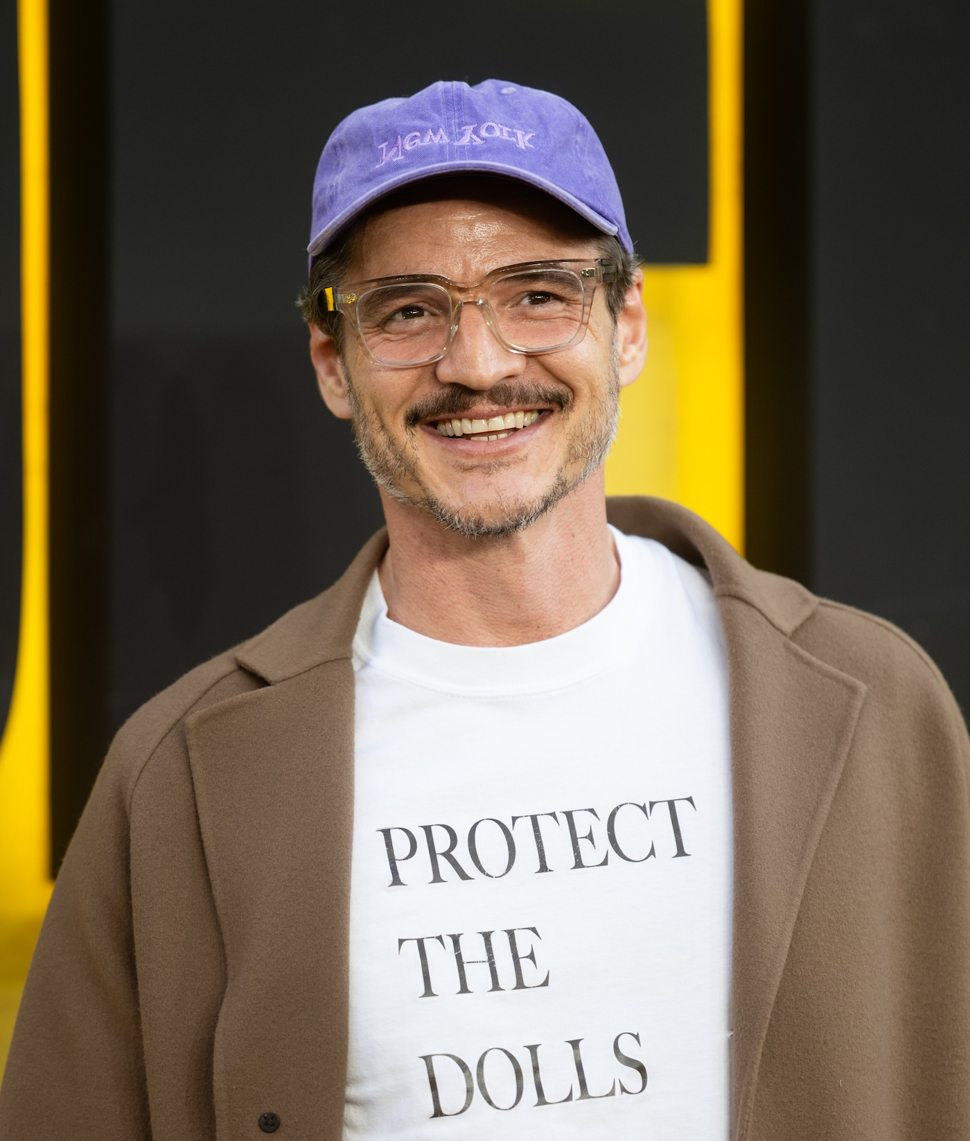 Pedro Pascal pozuje na czerwonym dywanie podczas specjalnego pokaz filmu „Thunderbolts*” w Londynie, ma na sobie biały T-shirt z nadrukiem „Protect the Dolls” projektu Connera Ivesa, długi, jasnobrązowy płaszcz  i fioletową czapkę z daszkiem.