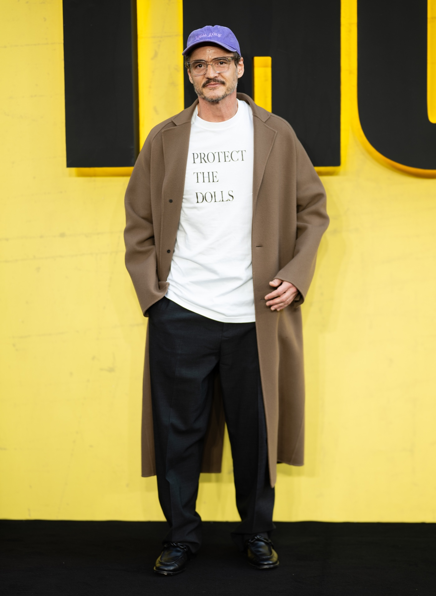 Pedro Pascal pozuje na czerwonym dywanie podczas specjalnego pokaz filmu „Thunderbolts*” w Londynie, ma na sobie biały T-shirt z nadrukiem „Protect the Dolls” projektu Connera Ivesa, długi, jasnobrązowy płaszcz, luźne, czarne spodnie zaprasowane w kant, czarne skórzane mokasyny i fioletową czapkę z daszkiem.