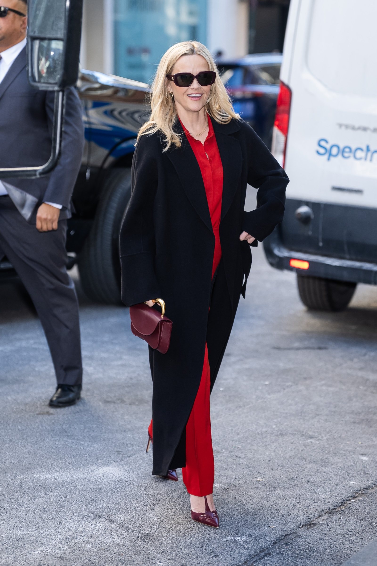Reese Witherspoon w czarnej długiej kurteczce, czerwona koszula damska, czerwone spodnie materiałowe, bordowe czółenka T-bar.