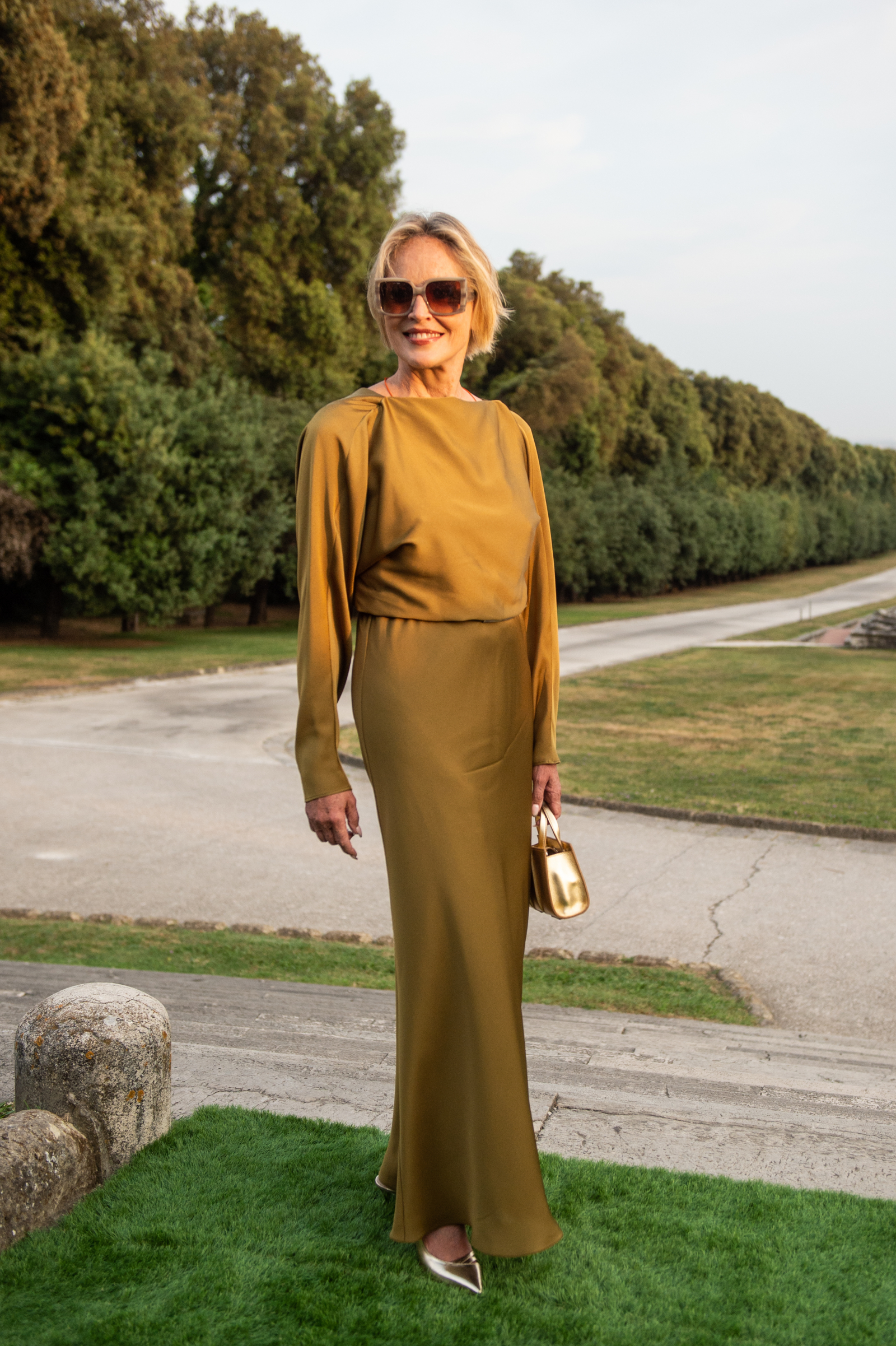 Sharon Stone w długiej sukience, trzyma złotą torebkę i nosi złote czółenka, pozując w plenerze.