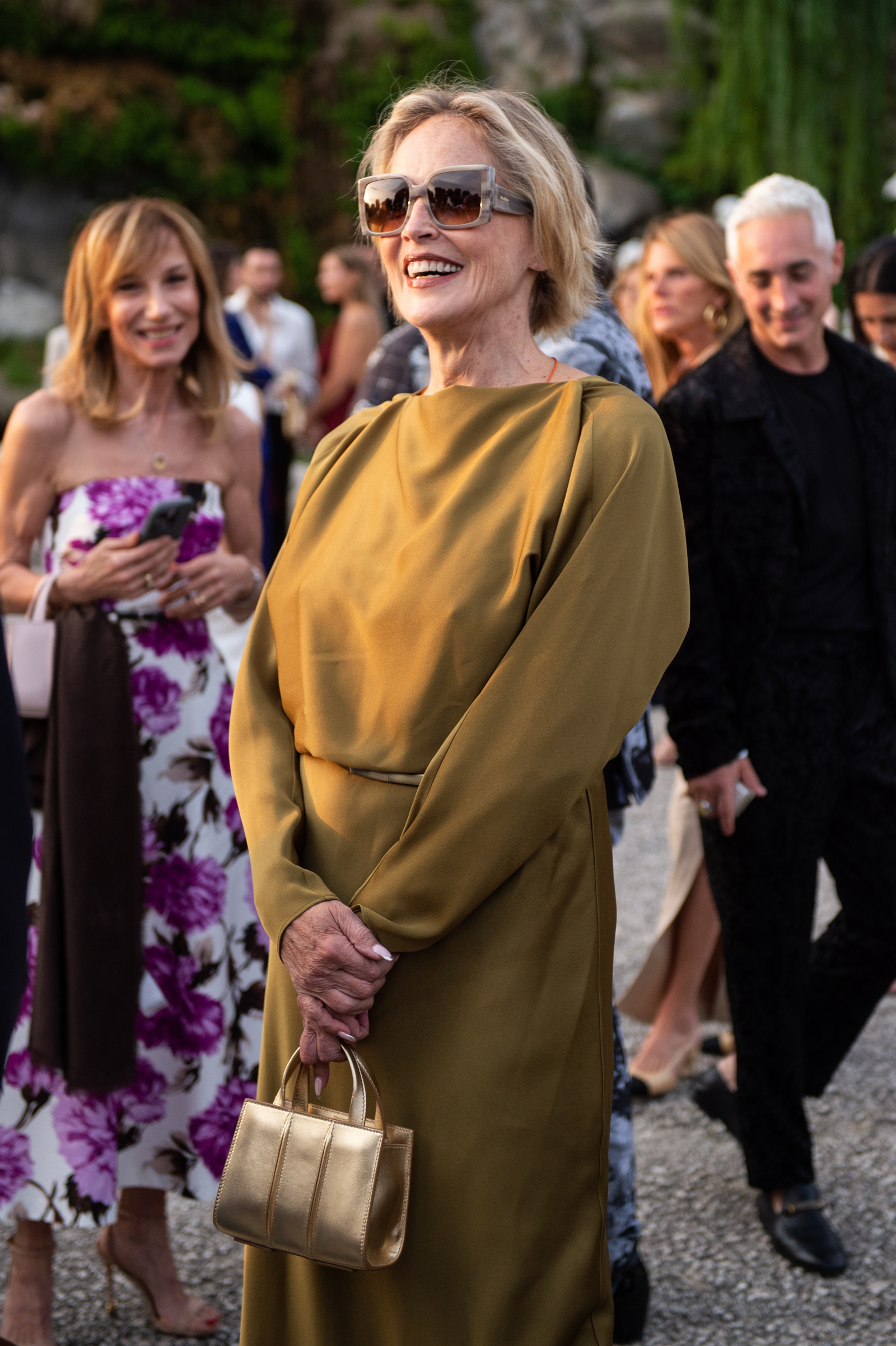 Sharon Stone w eleganckiej sukience, trzyma złotą torebkę, w okularach przeciwsłonecznych. W tle inni goście.