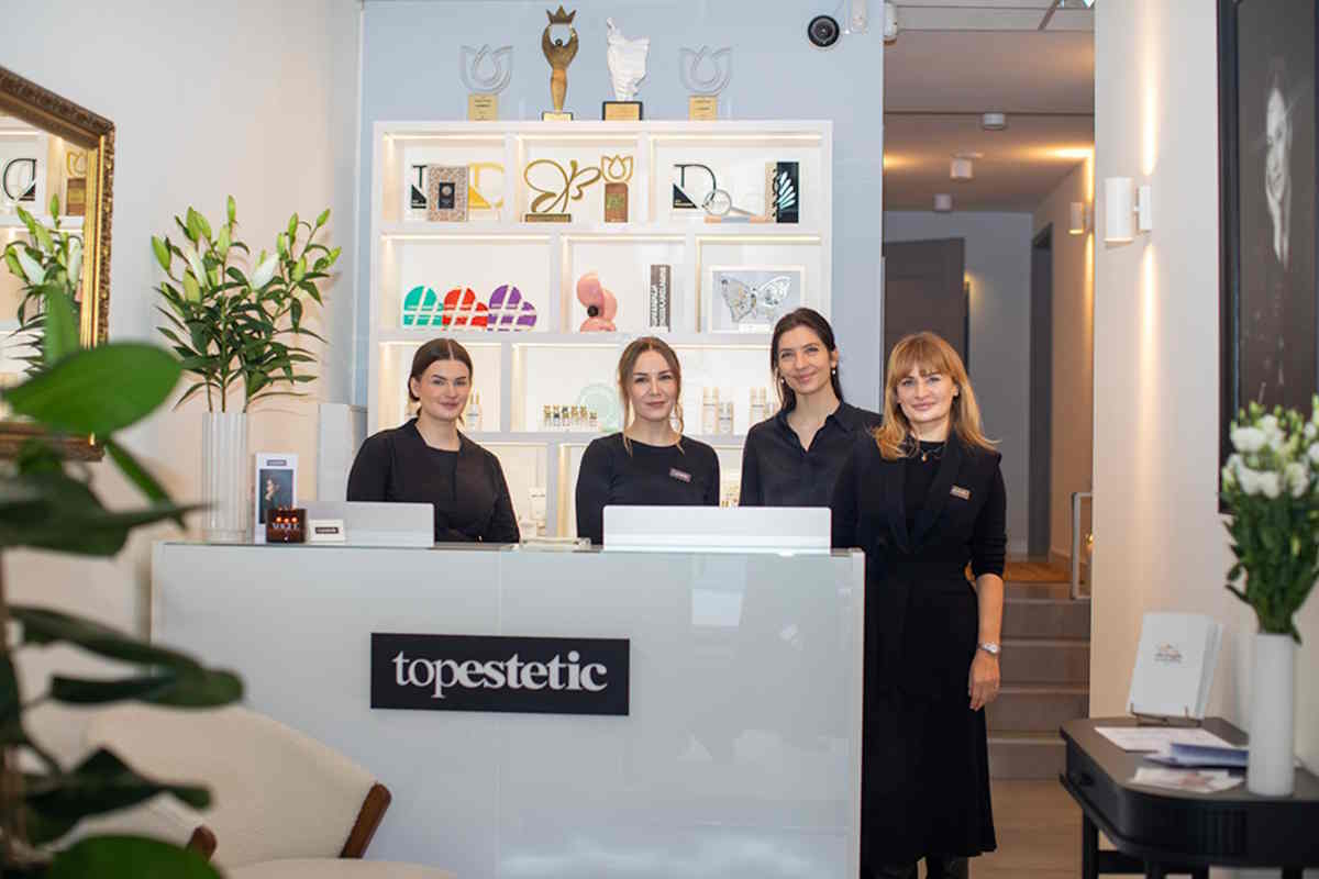 Cztery kobiety w uniformach stoją przy recepcji salonu kosmetycznego Topestetic, za nimi półka z produktami i trofeami.