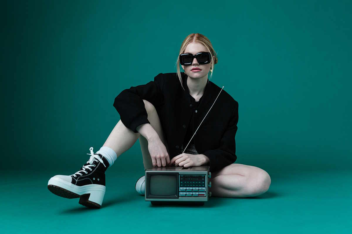 Kobieta w czarnych okularach przeciwsłonecznych siedzi na podłodze z radiem, ubrana w czarną kurtkę, białe skarpetki i sneakersy Converse na obcasie.