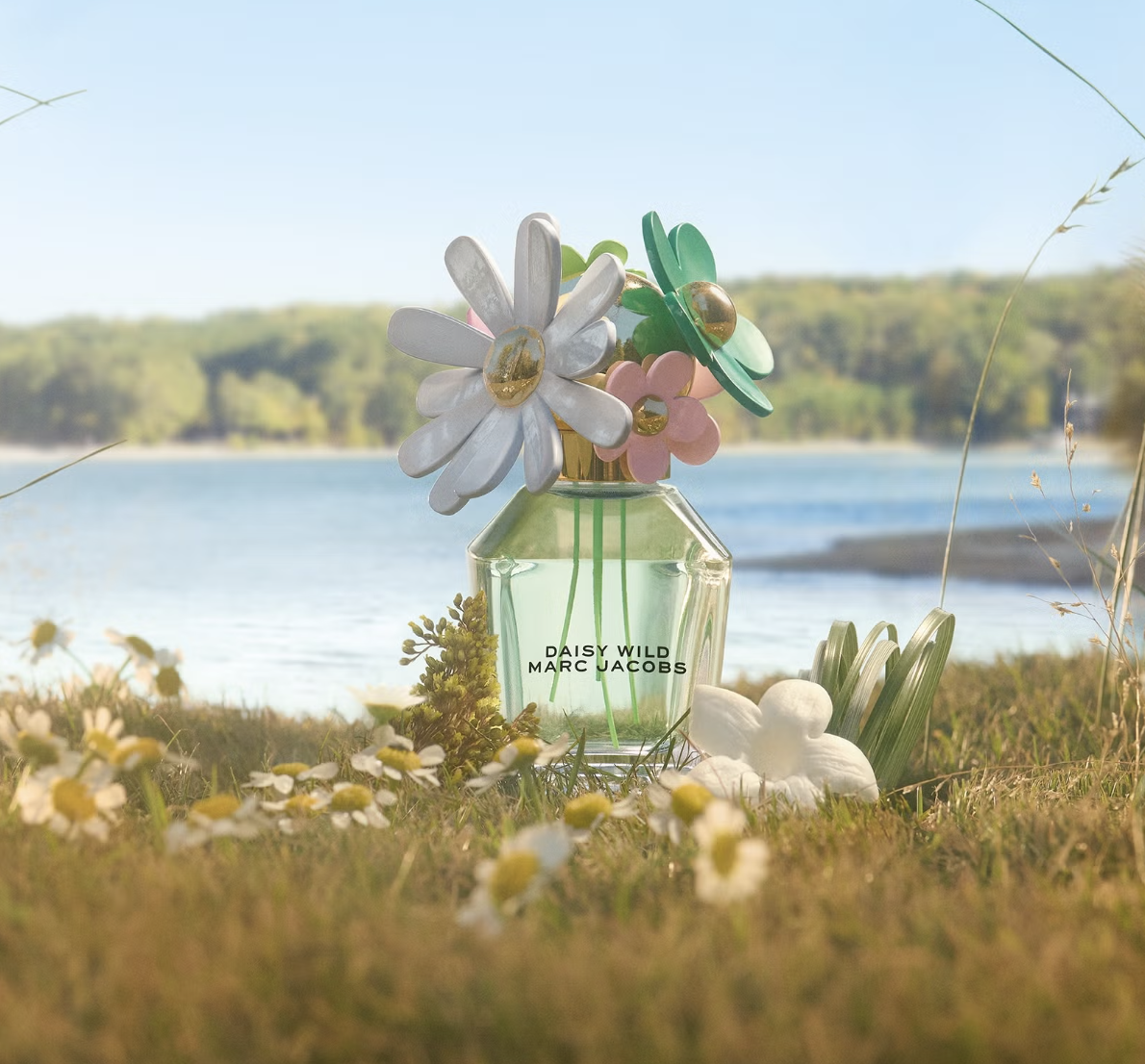 Butelka perfum Daisy Wild od Marc Jacobs z kolorowymi kwiatami na korku, na tle jeziora i zielonej trawy.