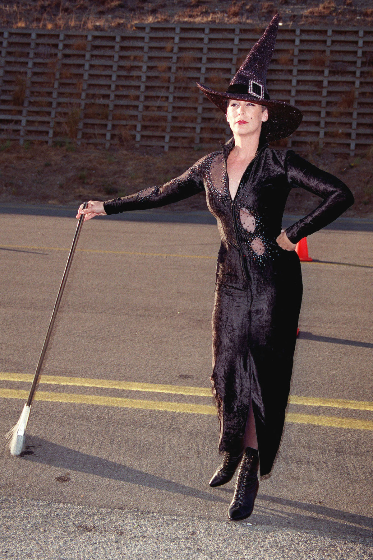 Kobieta w czarnej sukni i kapeluszu czarownicy trzyma szczotkę, pozując na tle drogi z wyznaczonymi liniami.