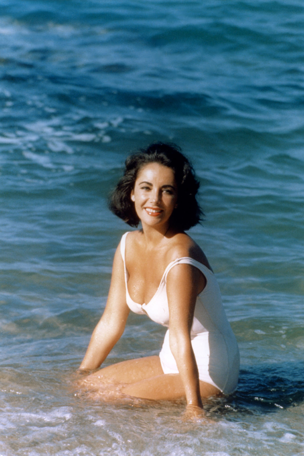 Elizabeth Taylor w białym kostiumie kąpielowym, siedząca na plaży nad morzem.