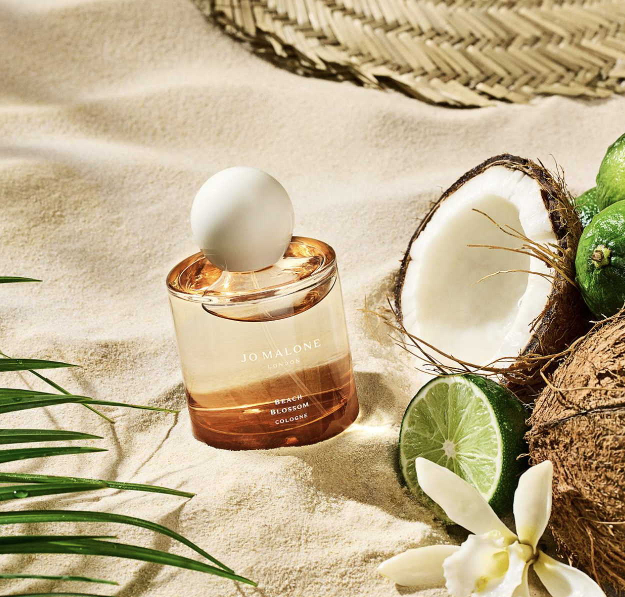 Perfumy pachnące jak dzień na plaży: Na piasku leży flakon perfum Jo Malone z napisem "Beach Blossom", obok kokosa, limonki i kwiatów.