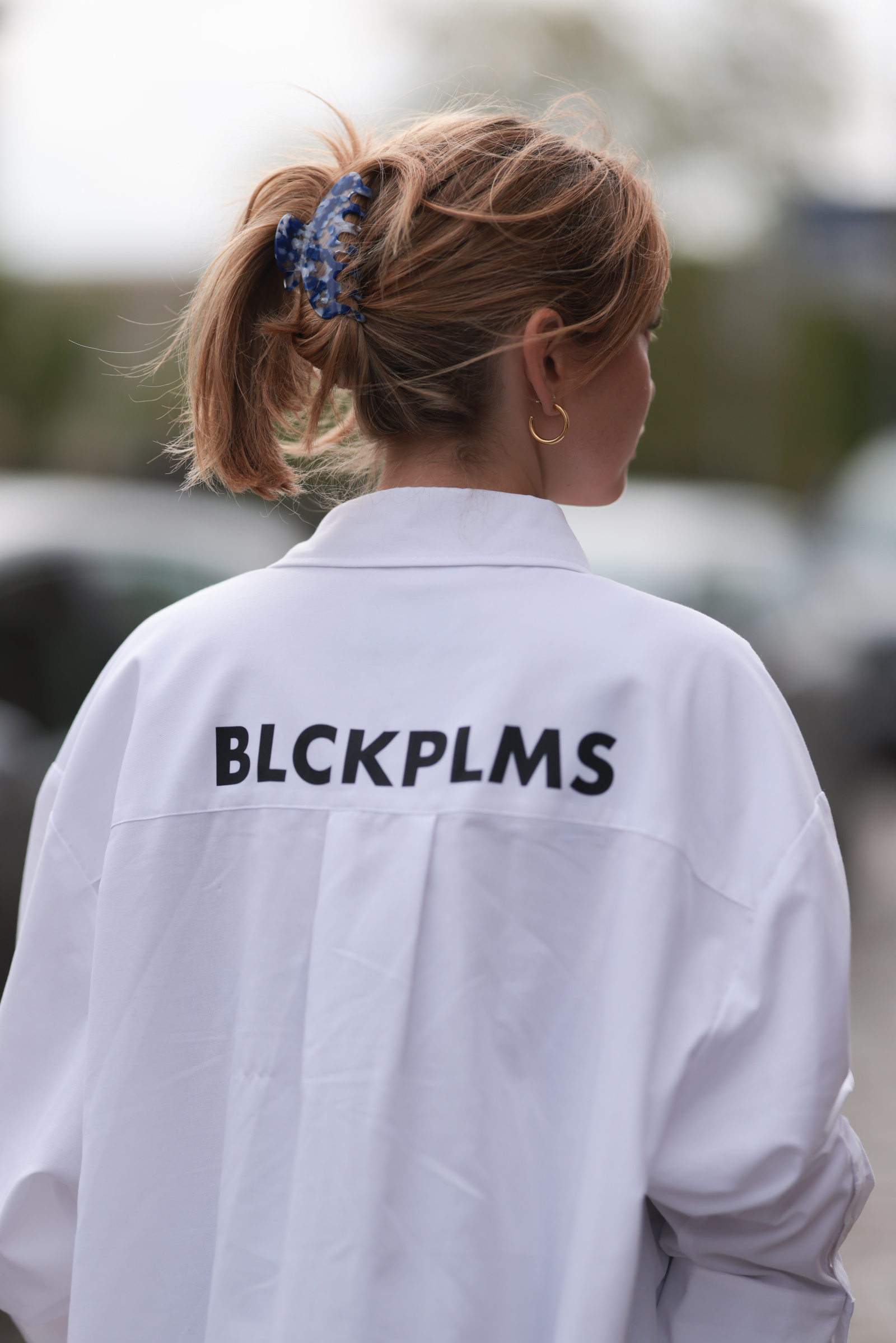 Kobieta z krótkimi włosami związanymi w kucyk, ubrana w białą koszulę z napisem "BLCKPLMS" z tyłu.