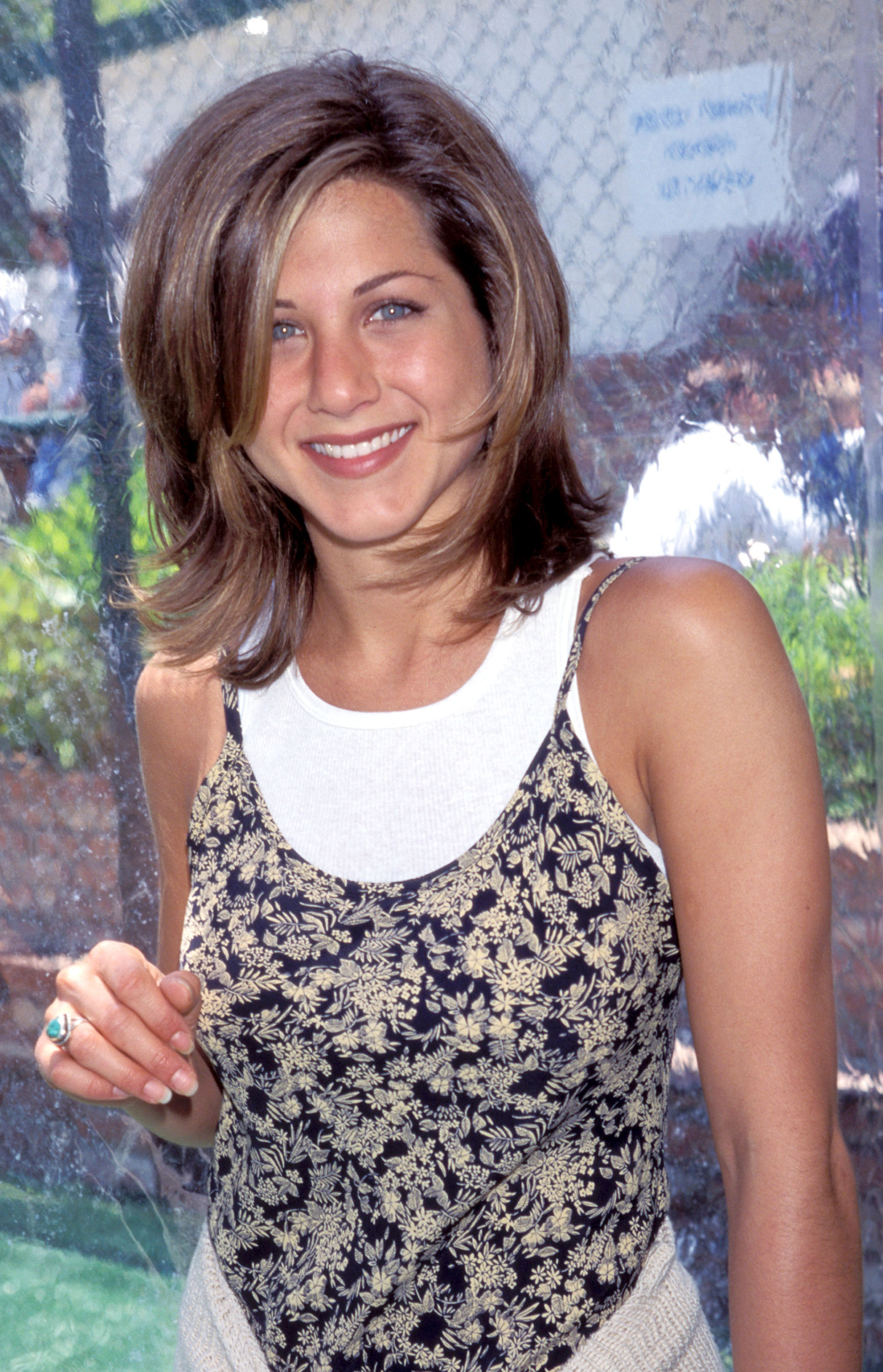 Jennifer Aniston uśmiecha się w stylizacji z lat 90., ma usta w odcieniu nude, w kwiecistej bluzce.