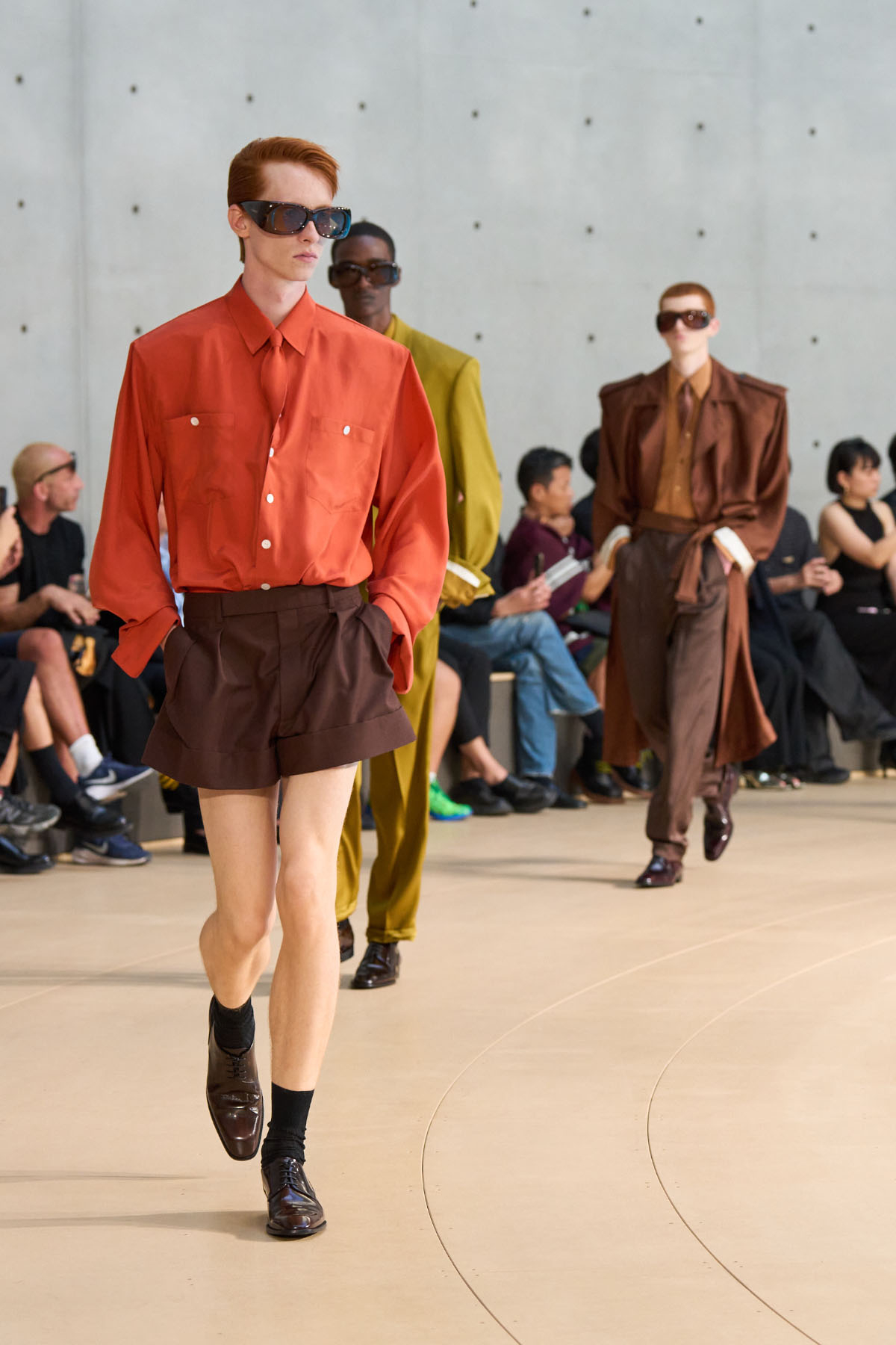 Model w stylowym pomarańczowym zestawie Saint Laurent na sezon wiosna-lato 2026 koszuli i brązowych szortach, na wybiegu obok innych modeli w eleganckich strojach. W tle widownia.