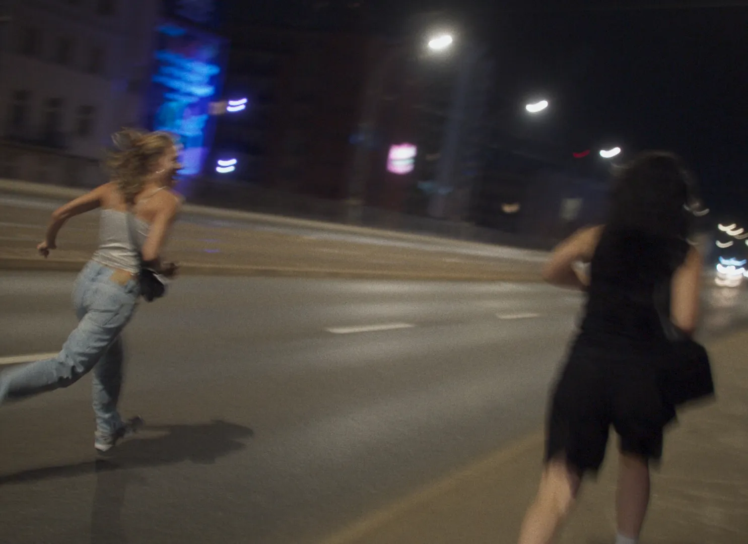 Lena Góra i Charli XCX uciekają w nocy, kadr z filmu "Erupcja". Dynamiczny ruch i miejskie światła w tle.