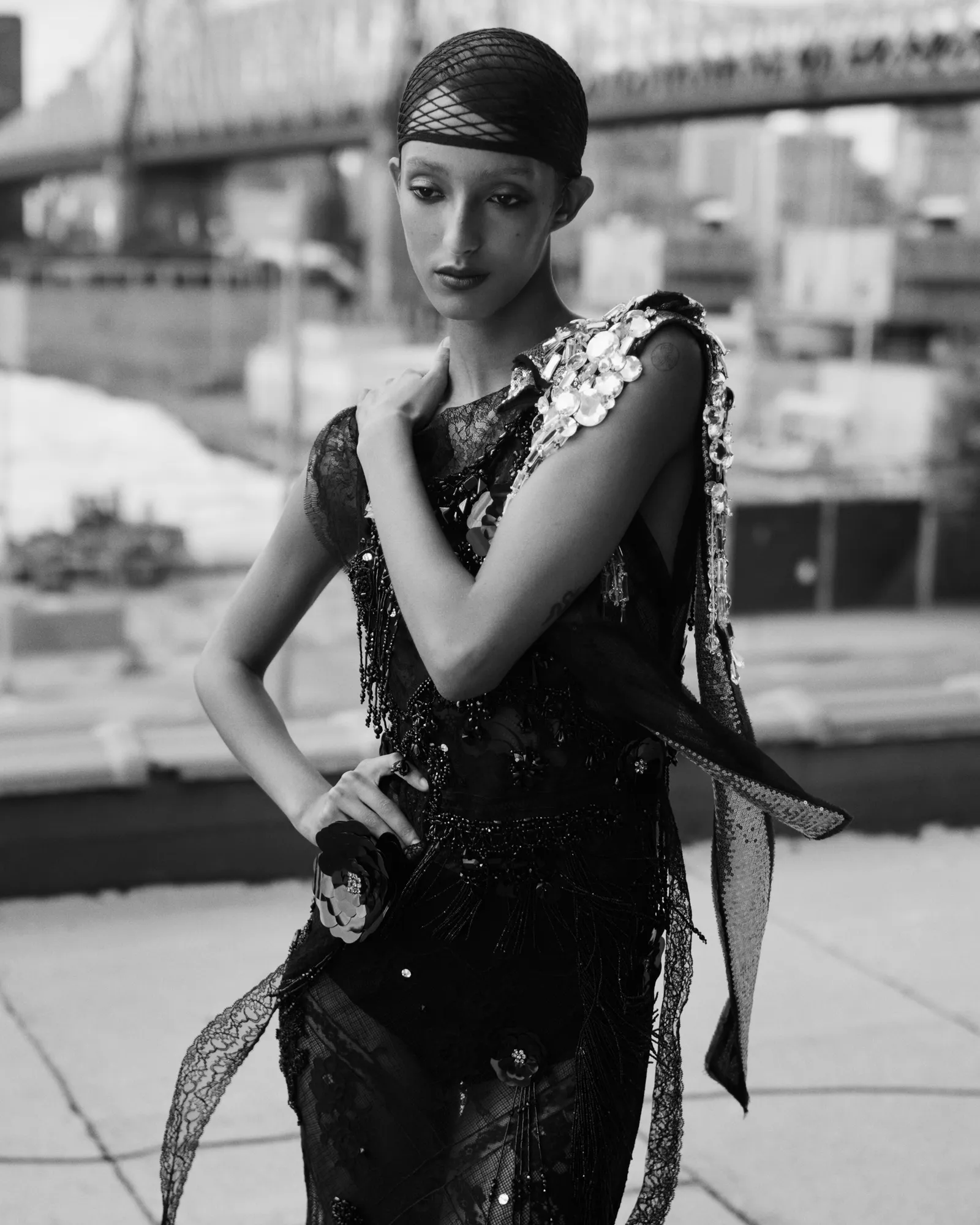 Modelka w eleganckiej, czarnej kreacji z detalami, pozująca na tle miejskiego krajobrazu. Artystyczne ujęcie w monochromatycznych odcieniach.