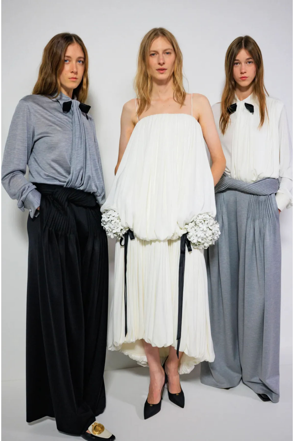 Trzy modelki w eleganckich stylizacjach Christian Dior. Jedna w białej sukience z dodatkami, pozostałe w stonowanych bluzkach i obszernych spodniach.