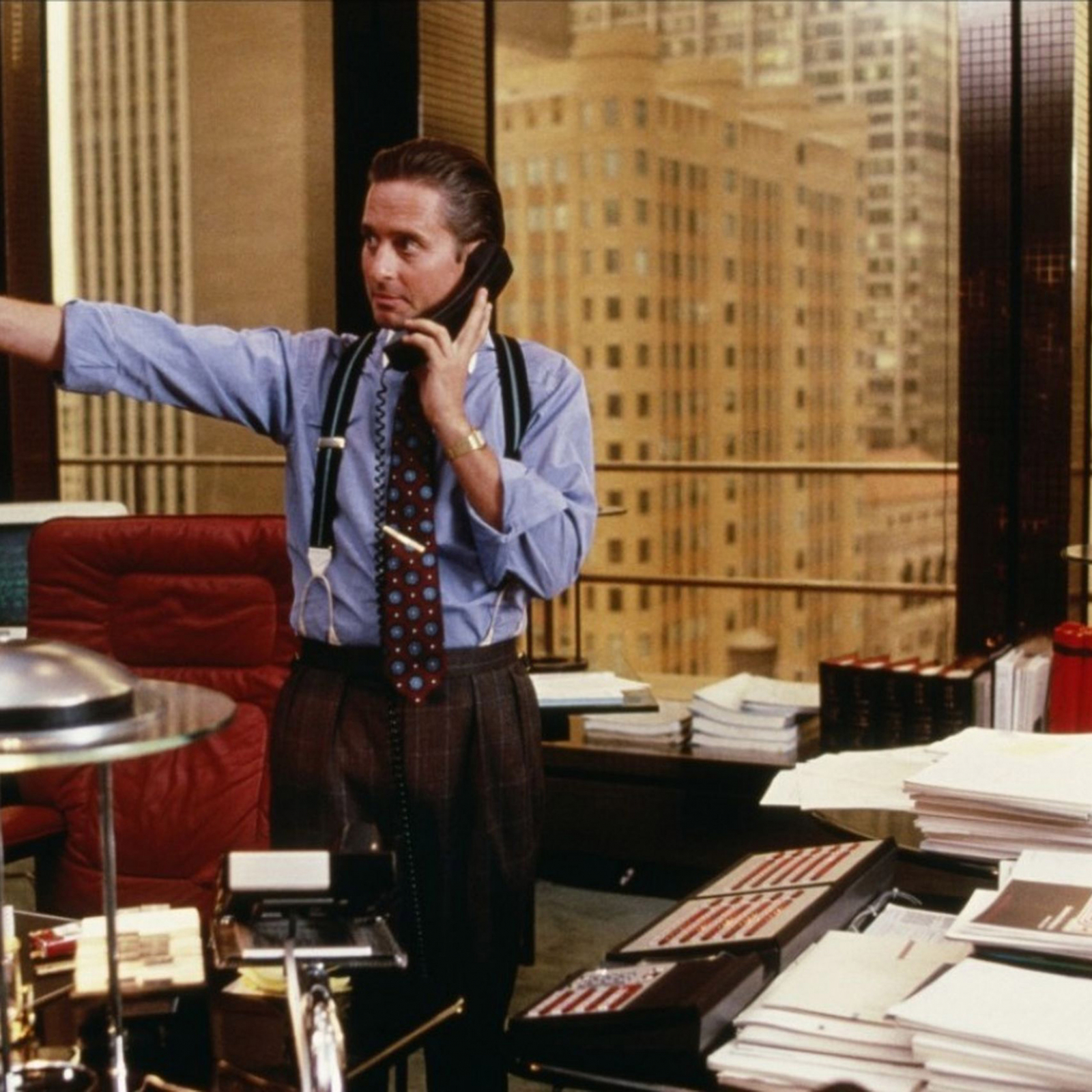 Michael Douglas w filmie "Wall Street", wykonujący telefon, w biurze z nowoczesnym widokiem na miasto.