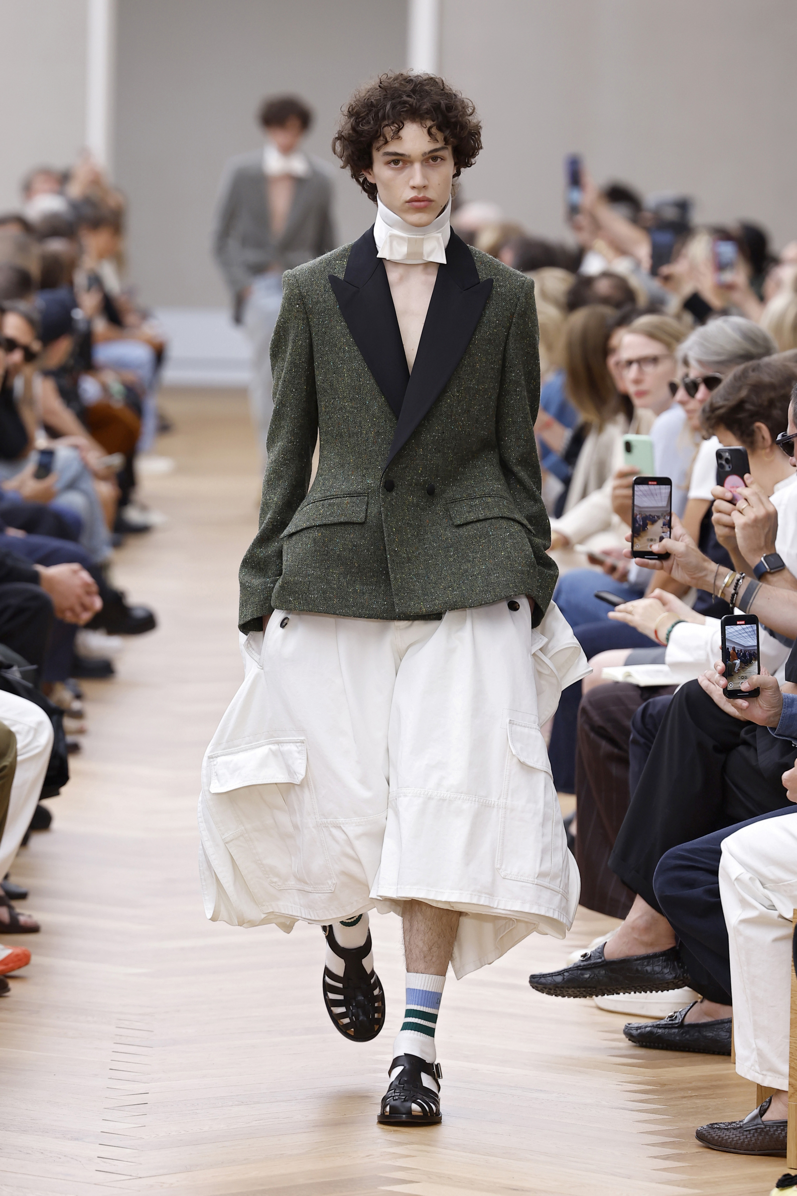 Pokaz mody męskiej Dior projektu Jonathana Andersona. Model w eleganckim żakiecie i luźnych spodniach idzie po wybiegu.