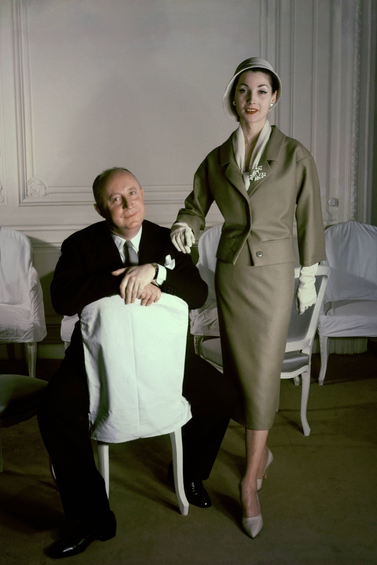 Christian Dior z modelką w eleganckiej kreacji, pozują przed białymi krzesłami w stylowym wnętrzu.