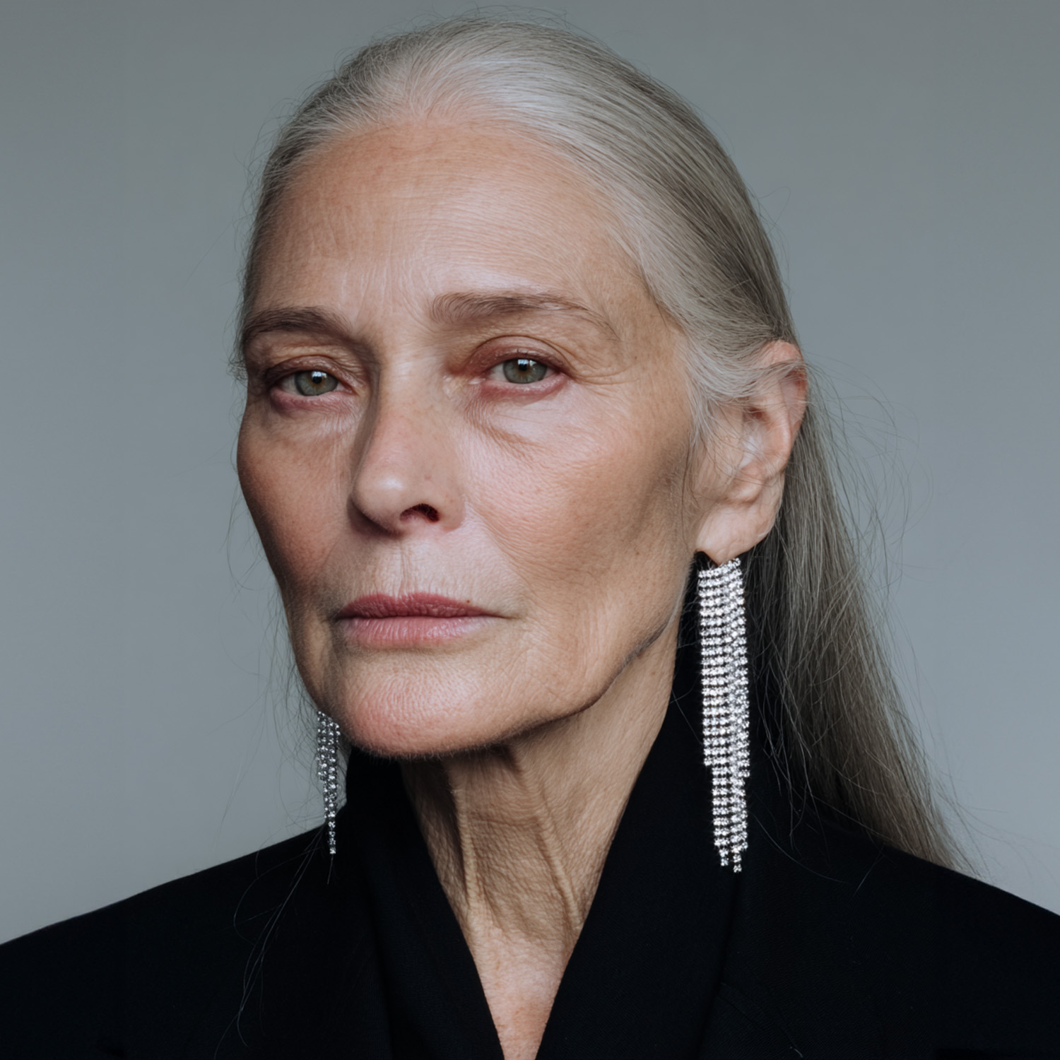 Portret dojrzałej kobiety z długimi siwymi włosami, w eleganckich kolczykach i czarnym płaszczu.