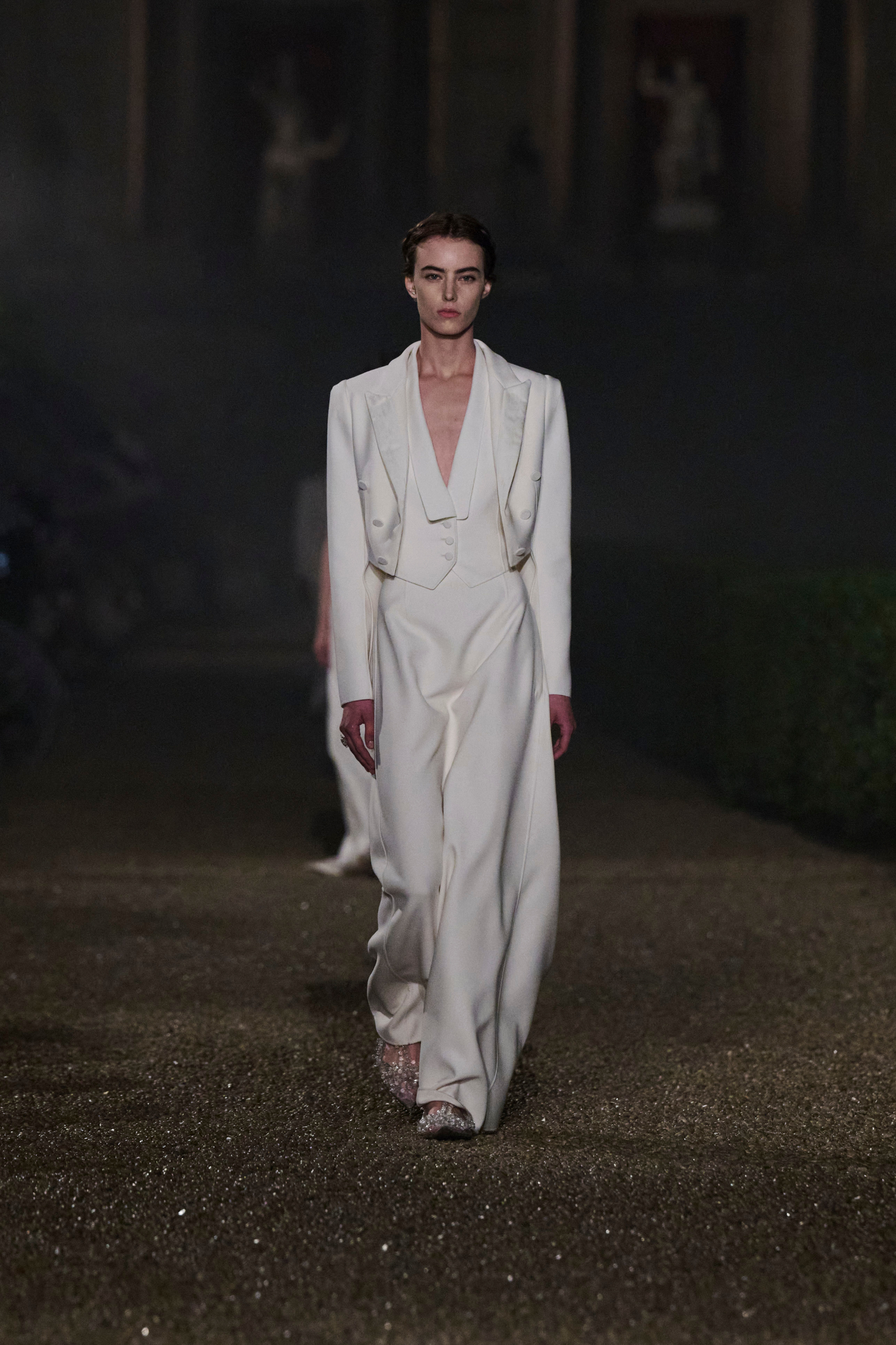 Modelka w białym eleganckim garniturze na wybiegu, w tle stylizowany ogród, lekka mgła.