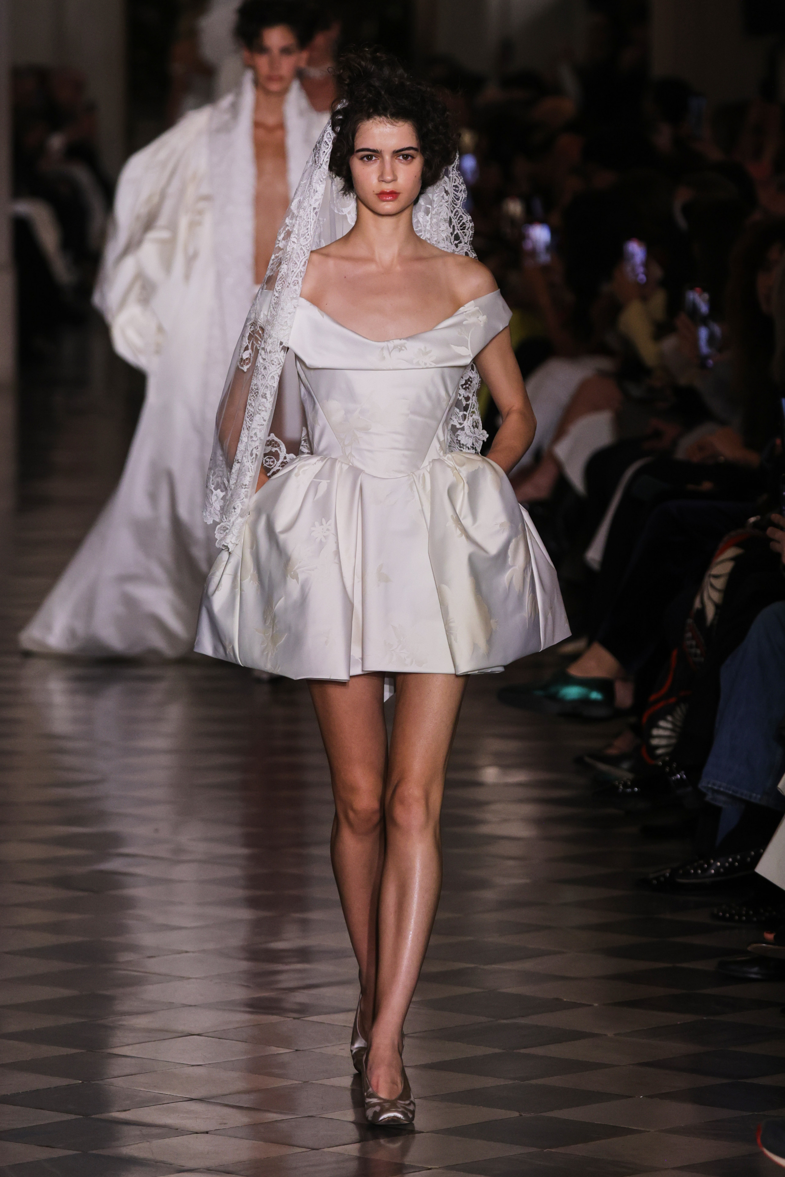 Modelka w białej, eleganckiej sukni Vivienne Westwood koronkowym welonem, idzie po wybiegu na pokazie mody. W tle widoczna publiczność.