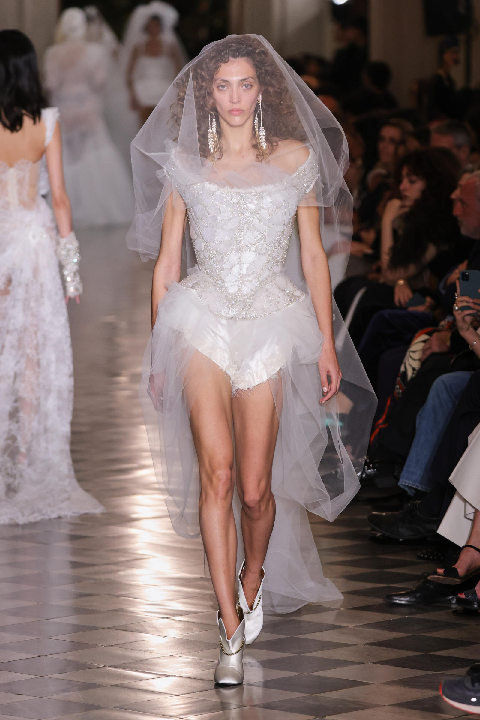 Modelka w kreacji Vivienne Westwood, w białym kostiumie z tiulowym welonem, idzie po wybiegu.