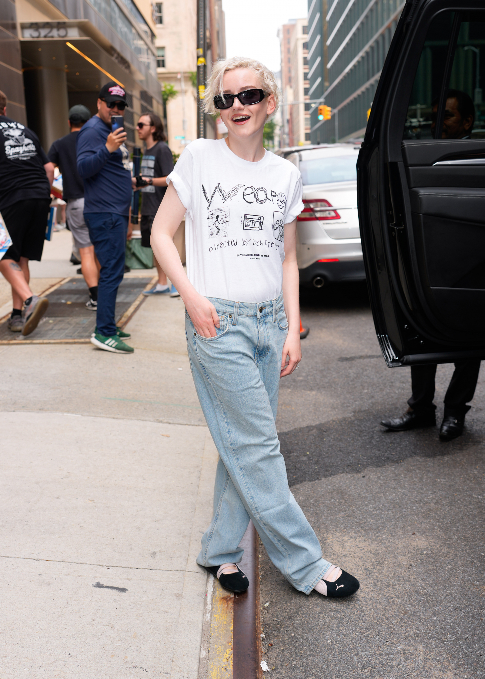 Julia Garner w białej koszulce i jeansach, nosi baleriny Puma Speedcat, uśmiechając się do kamery na ulicy.