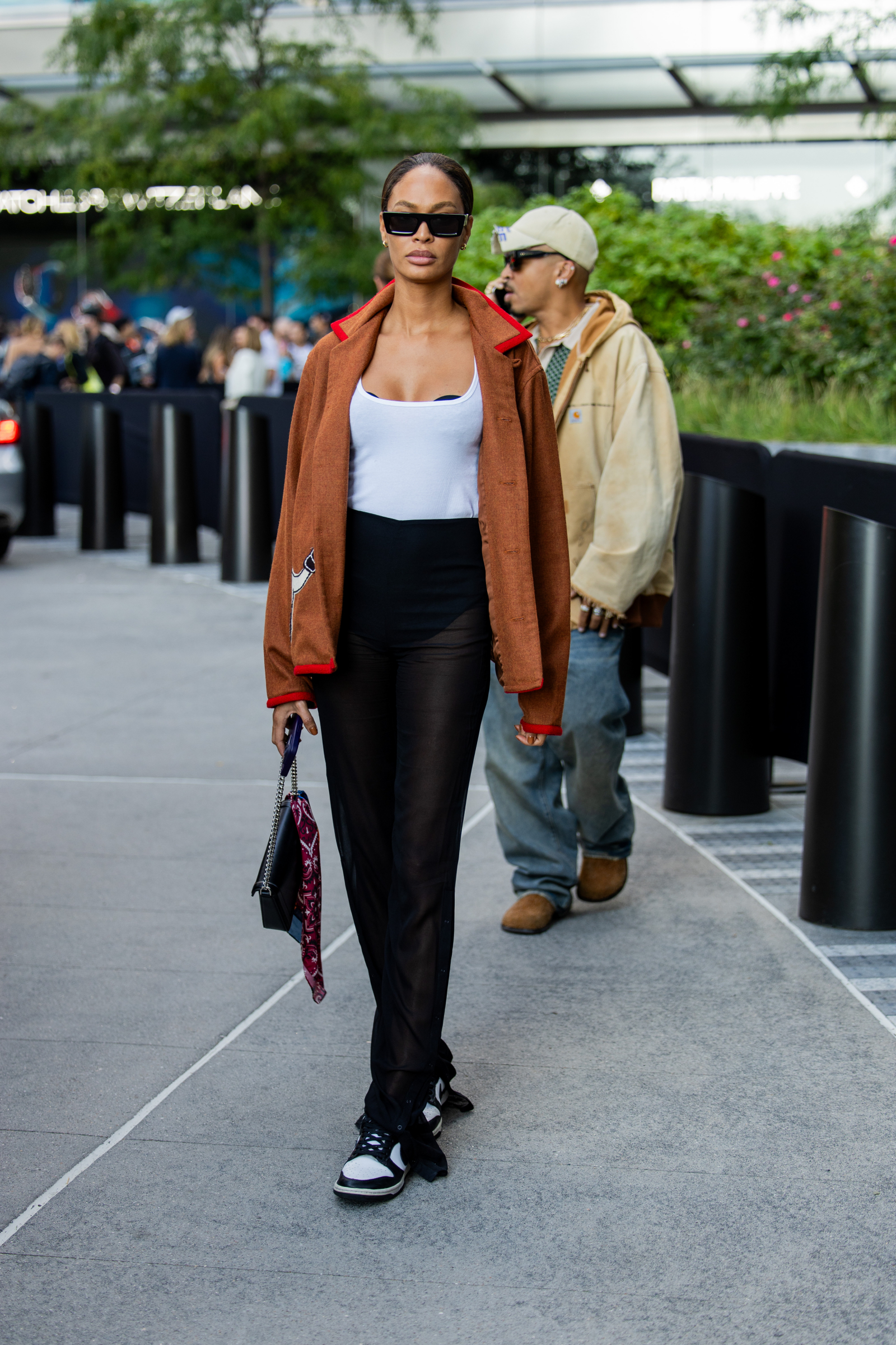 Joan Smalls w przezroczystych spodniach, białym topie i brązowej marynarce, nosi biało-czarne sneakersy Nike.